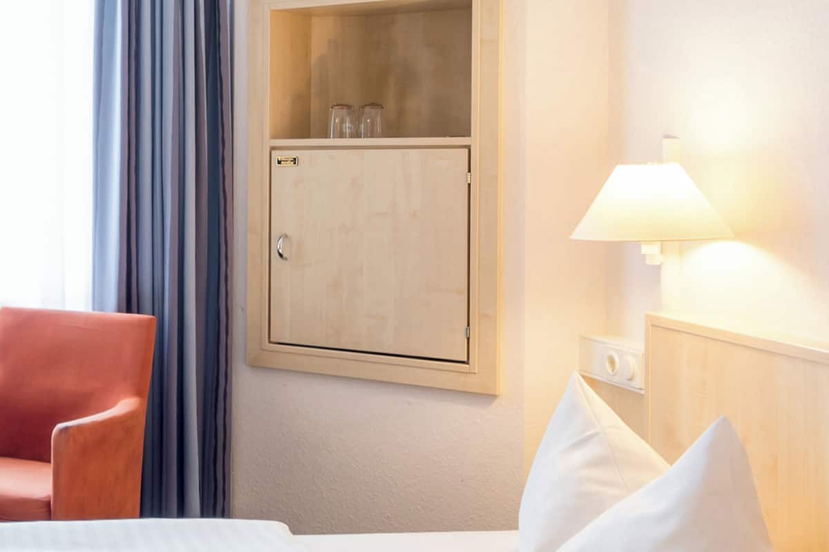 Ein Hotelzimmer mit einem Holzkoffer, einer Lampe, einem Stuhl und einem Bett mit Kissen.