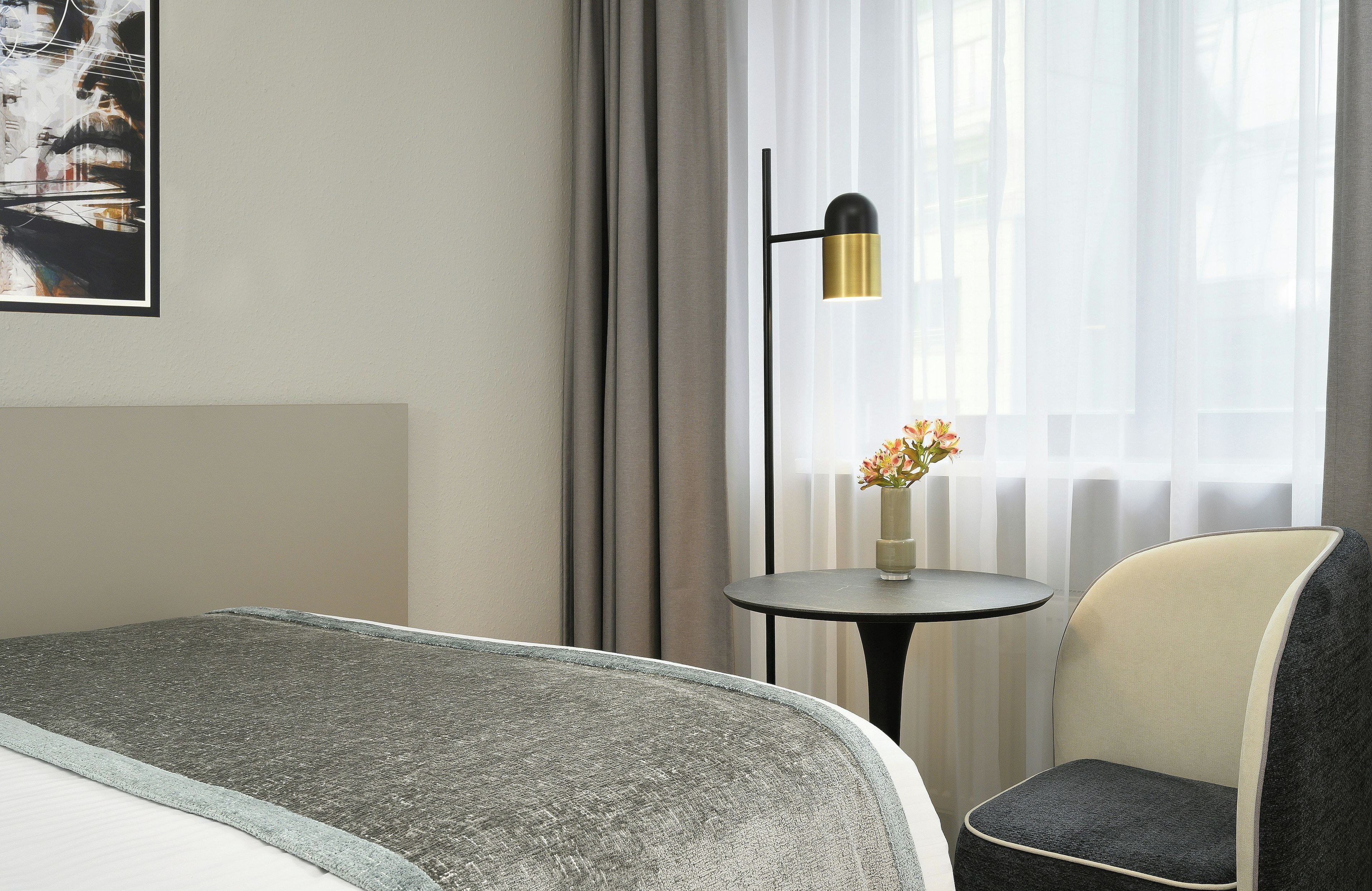 A hotel room with a bed, a chair, a small table with a vase of flowers, and a wall-mounted lamp.