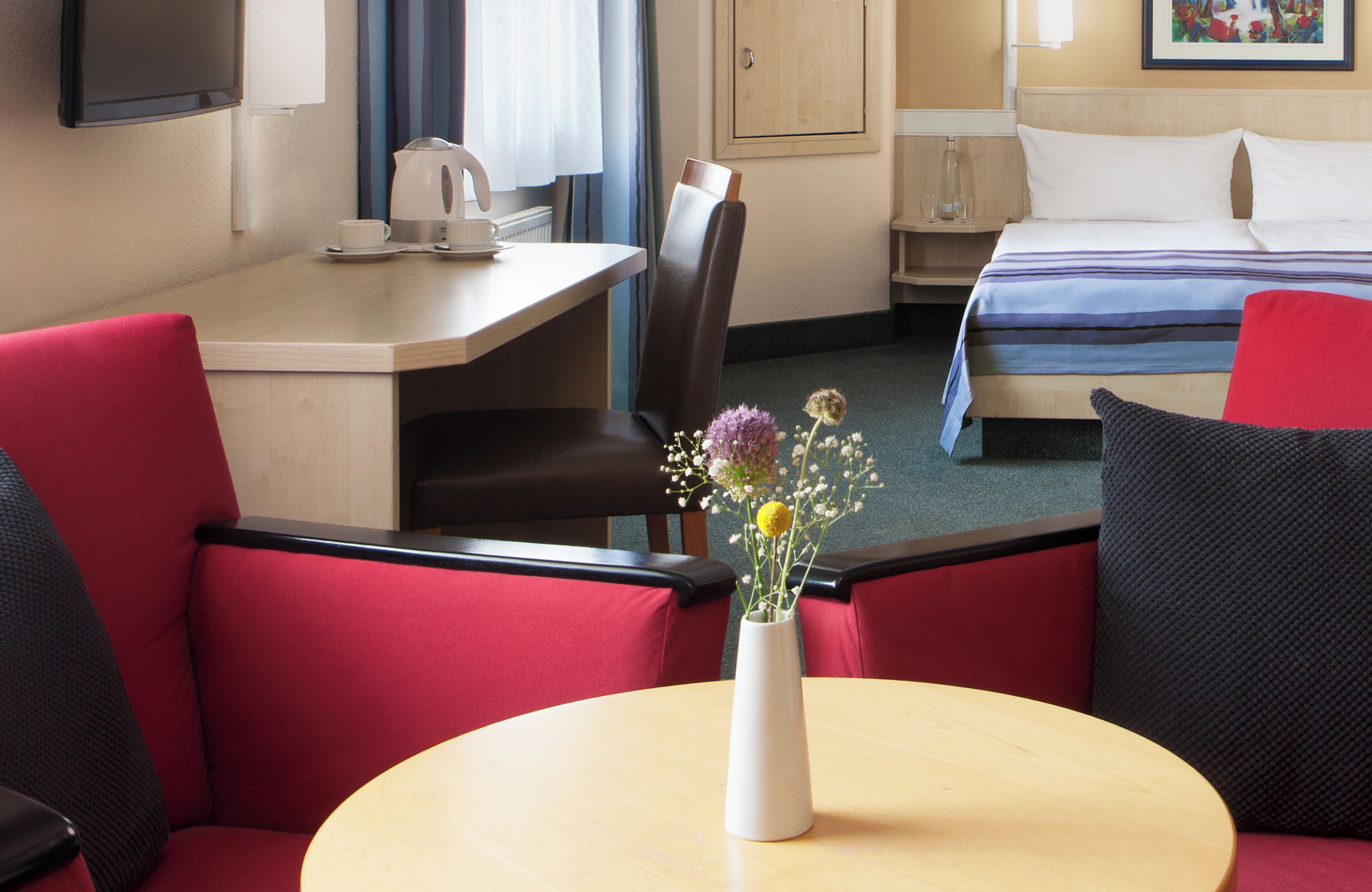 Habitación de hotel con cama, escritorio, silla, mesa con un jarrón de flores y televisor en la pared.