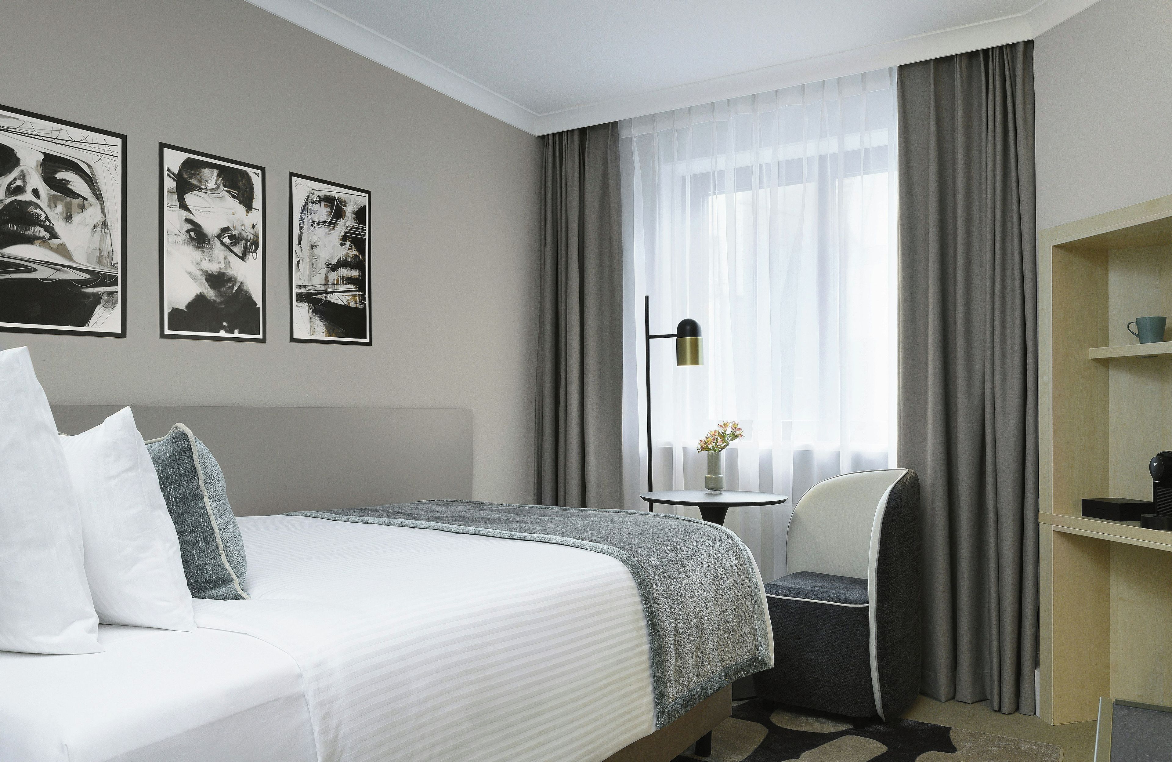 A hotel room with a bed, a chair, a small table, and a shelf. There are three framed pictures on the wall.