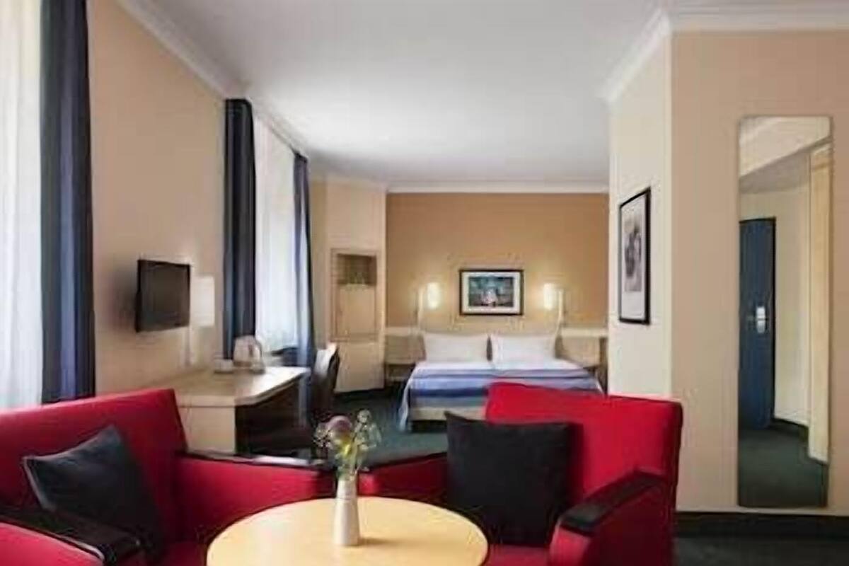A hotel room with a bed, a desk, a chair, a TV, and a small table with a vase.