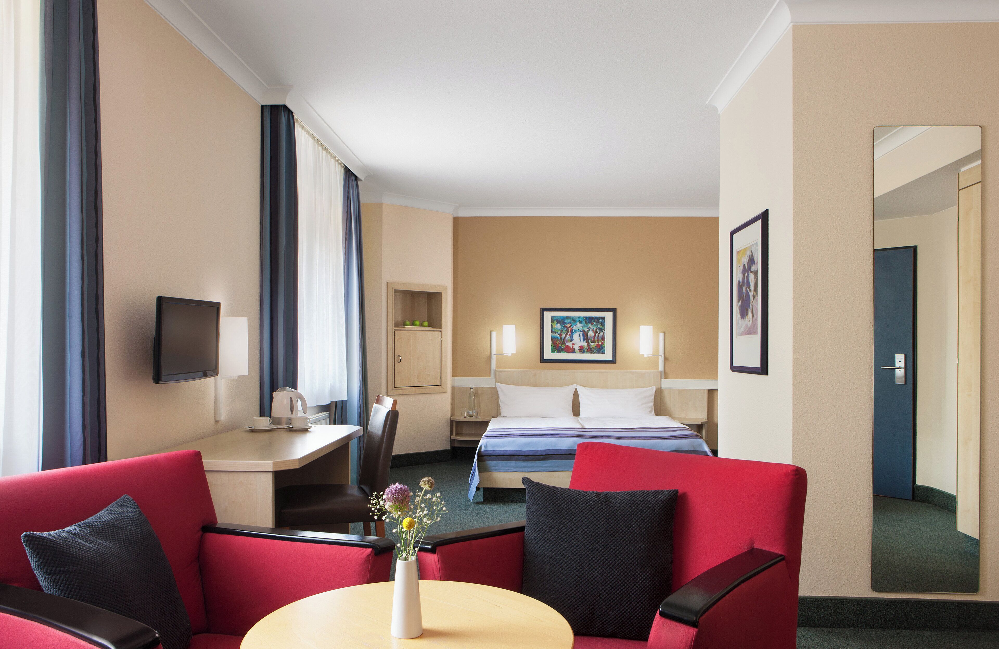 A hotel room with a bed, a desk with a chair, a TV, and a dining area with a table and chairs.