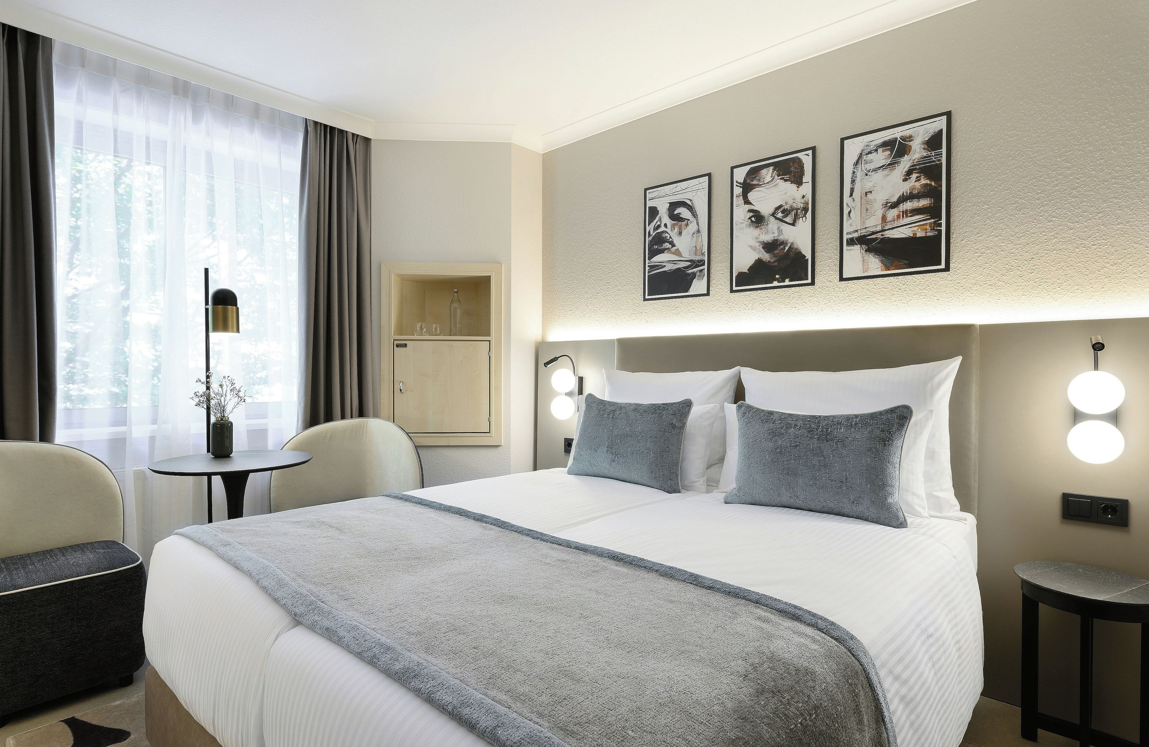 A hotel room with a bed, bedside tables, a desk, and a chair. There are wall-mounted lights and artwork above the bed.