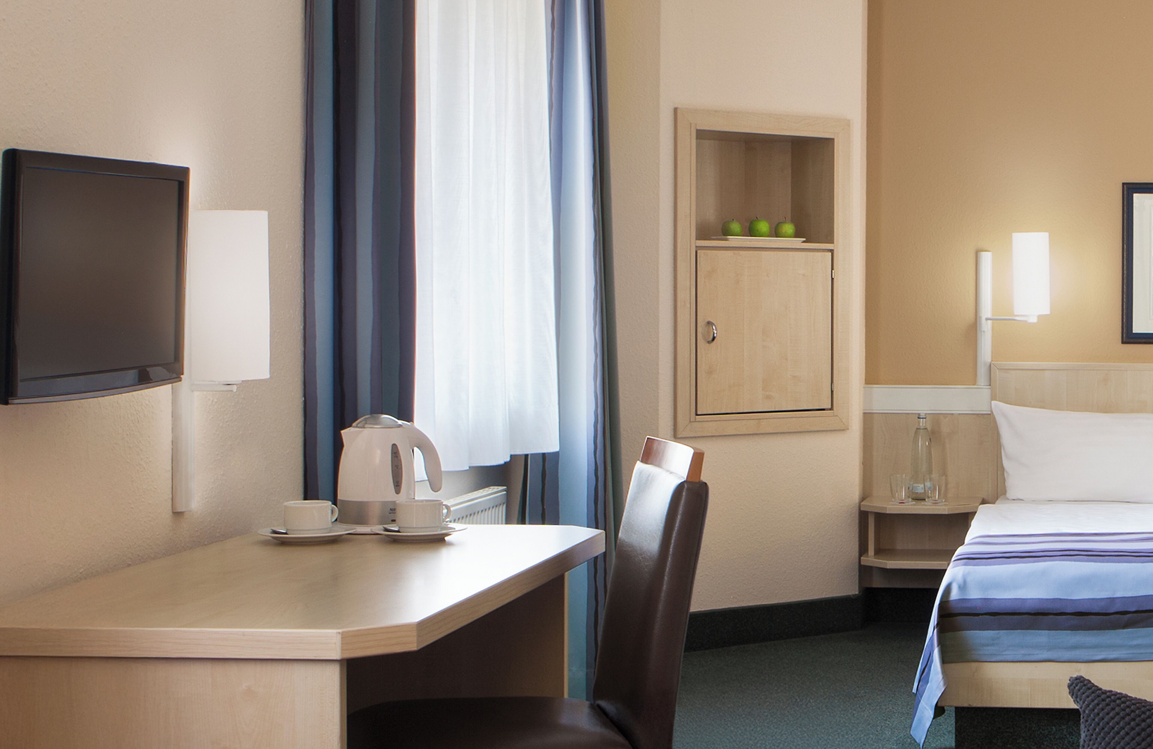 Habitación de hotel con cama, escritorio con cafetera, una silla, televisor, armario y mesita de noche con lámpara.
