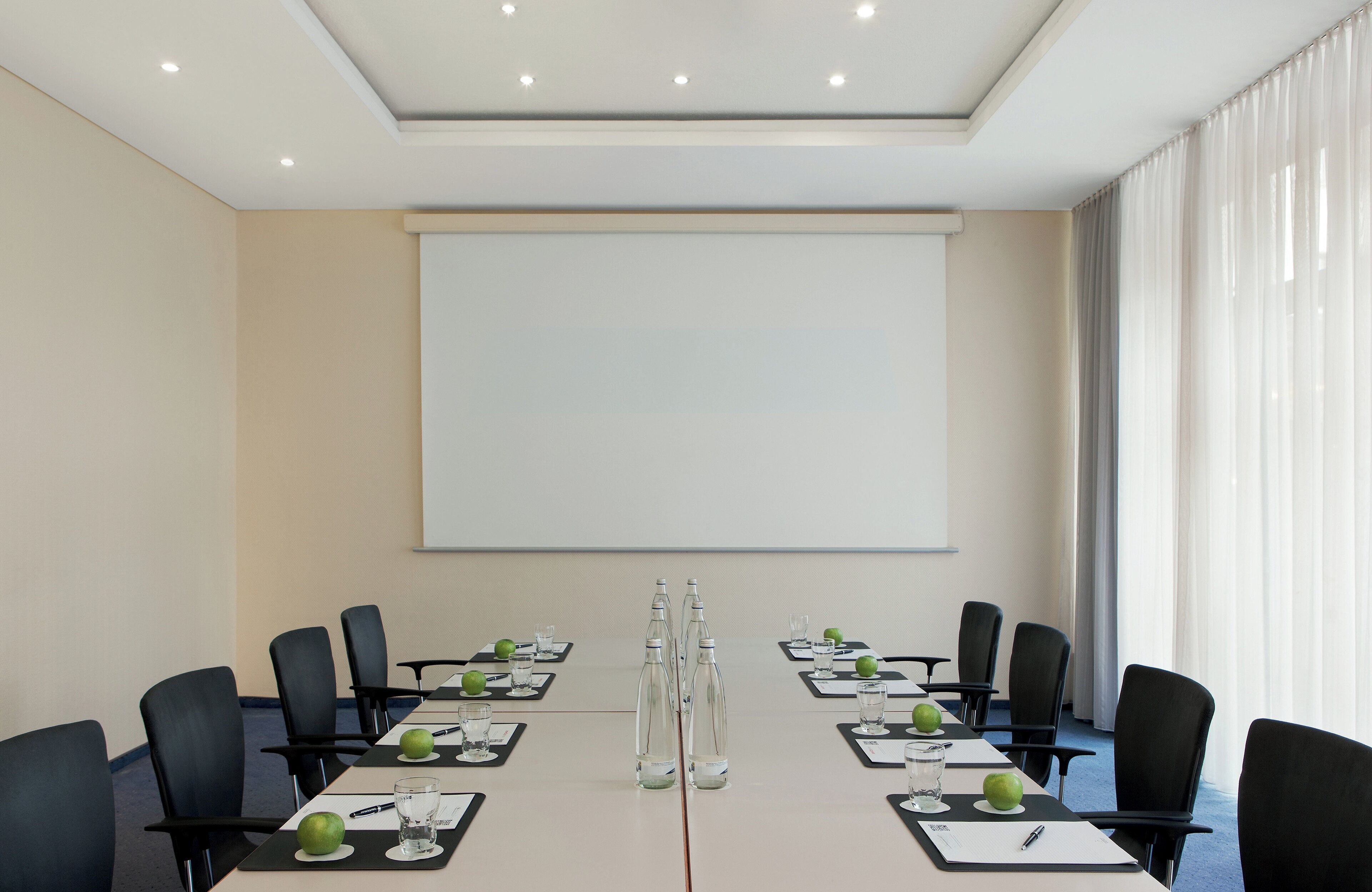 Un salón de conferencias con una mesa larga, sillas negras, botellas de agua y una pantalla de proyector.