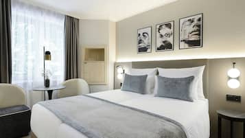 Habitación de hotel con cama, mesitas de noche, un escritorio y una silla. Hay luces fijadas en la pared y obras de arte sobre la cama.