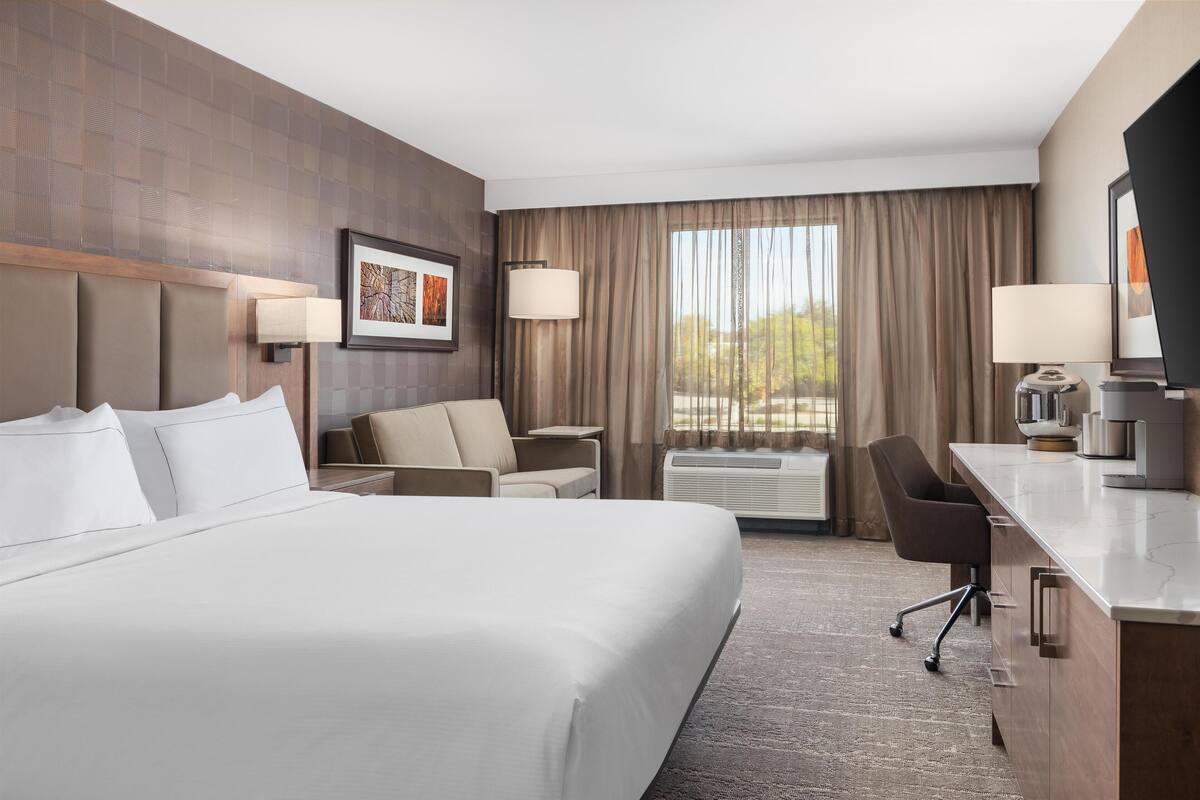 A hotel room with a large bed, a desk with a chair, a sofa, and a large window with curtains.