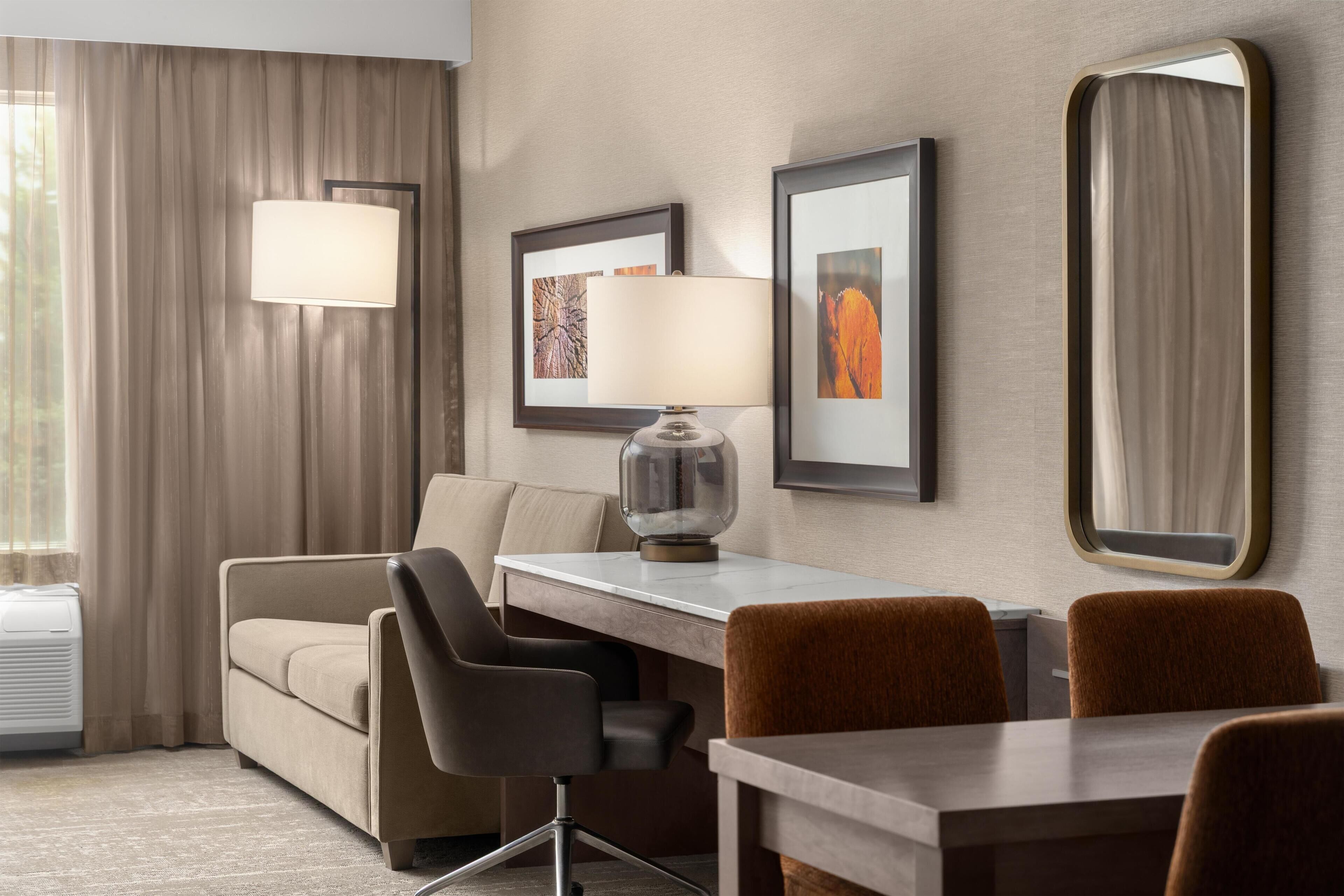 A hotel room with a sofa, two chairs, a dining table, a lamp, framed pictures, and a large mirror.