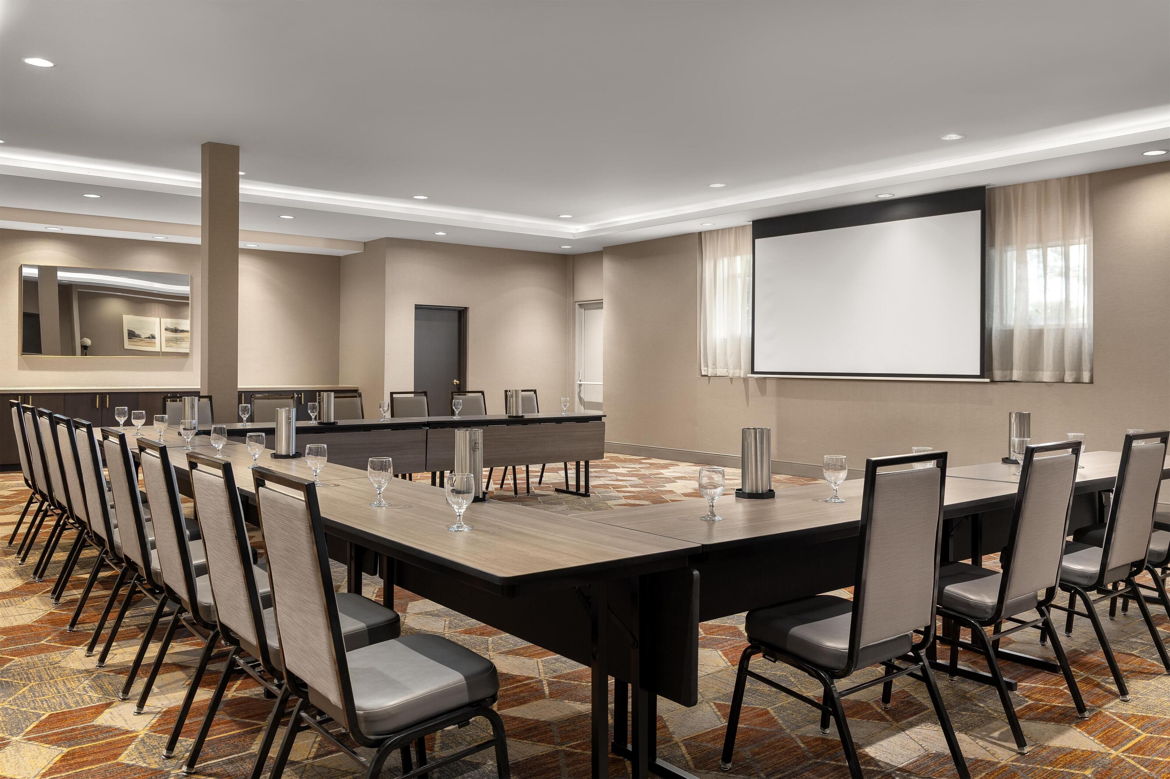 A conference room with a long table, chairs, a projector screen, and a large mirror.