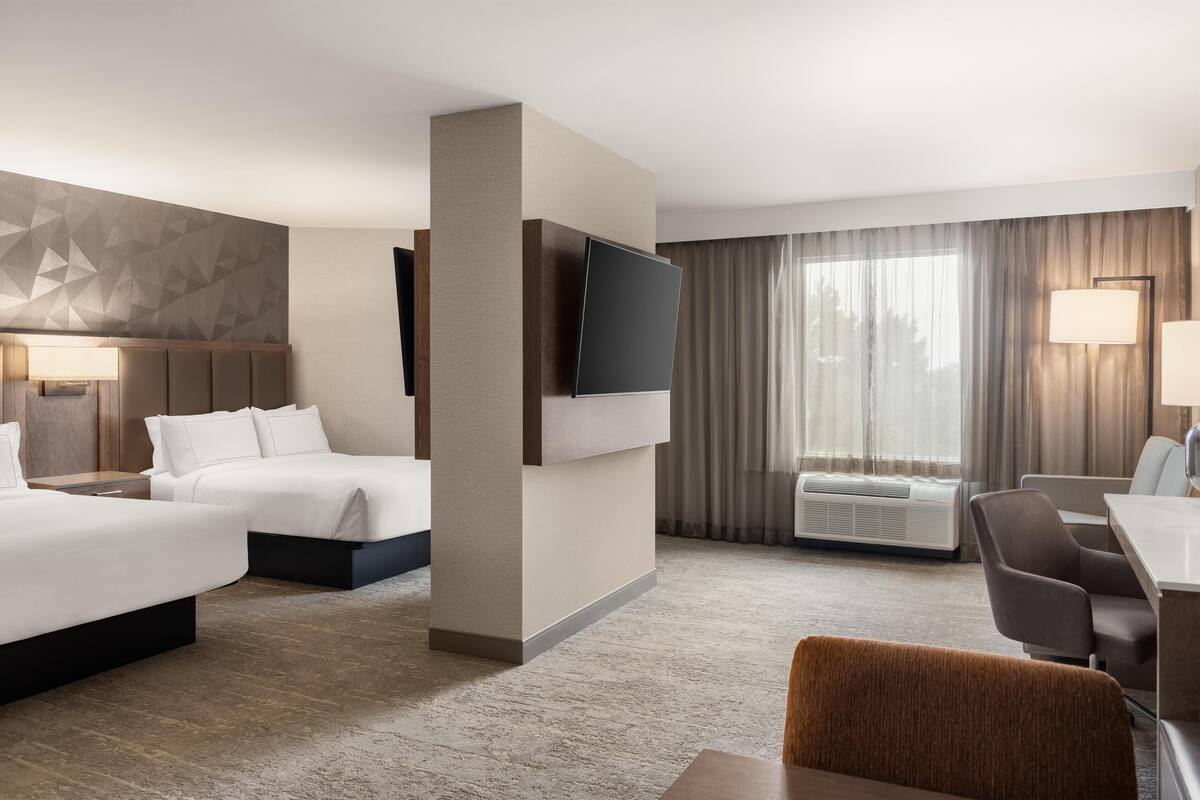 A hotel room with two beds, a flat-screen TV, a desk with a chair, and a large window with curtains.