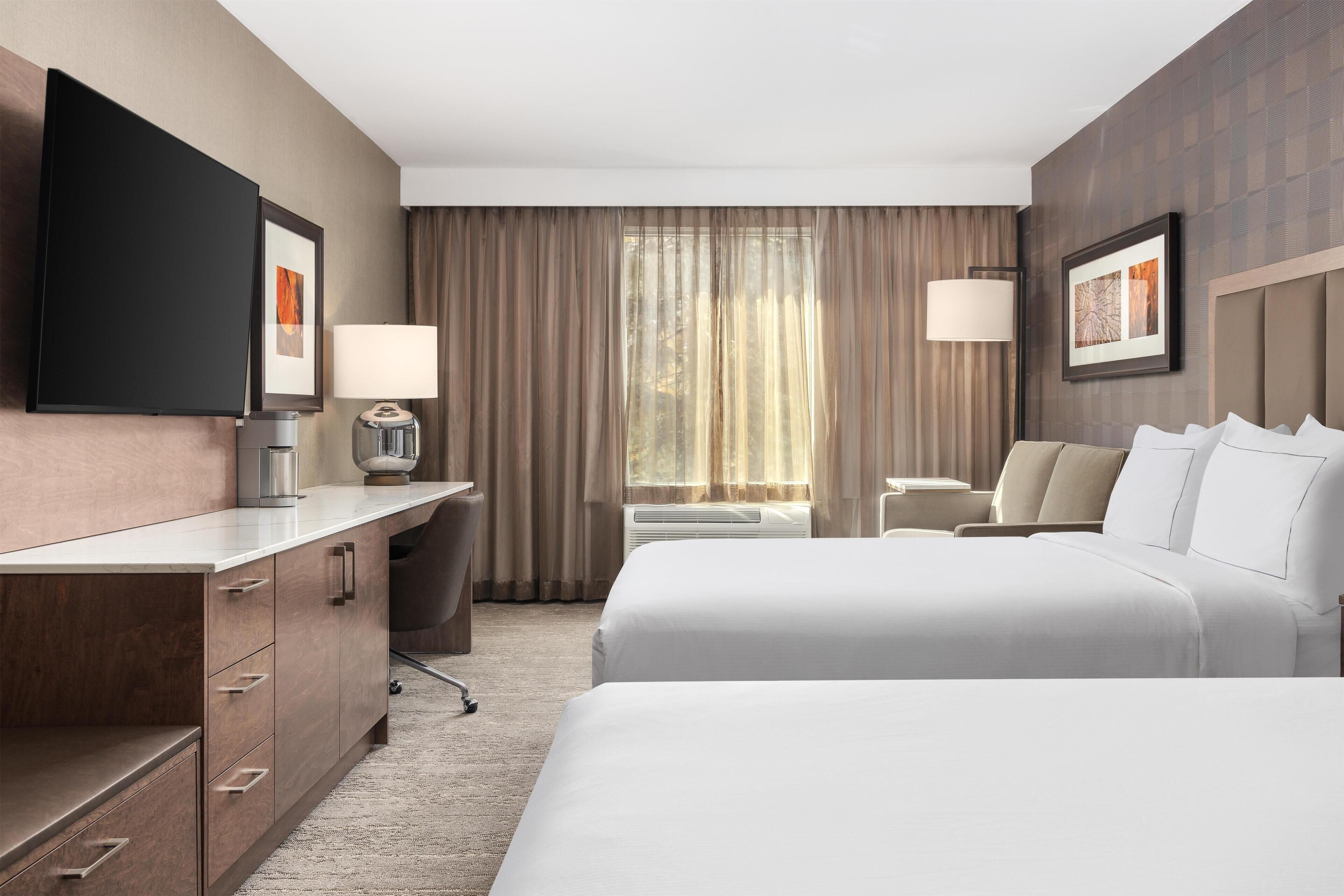 Een hotelkamer met twee bedden, een bureau, een stoel, een televisie en een raam met gordijnen.