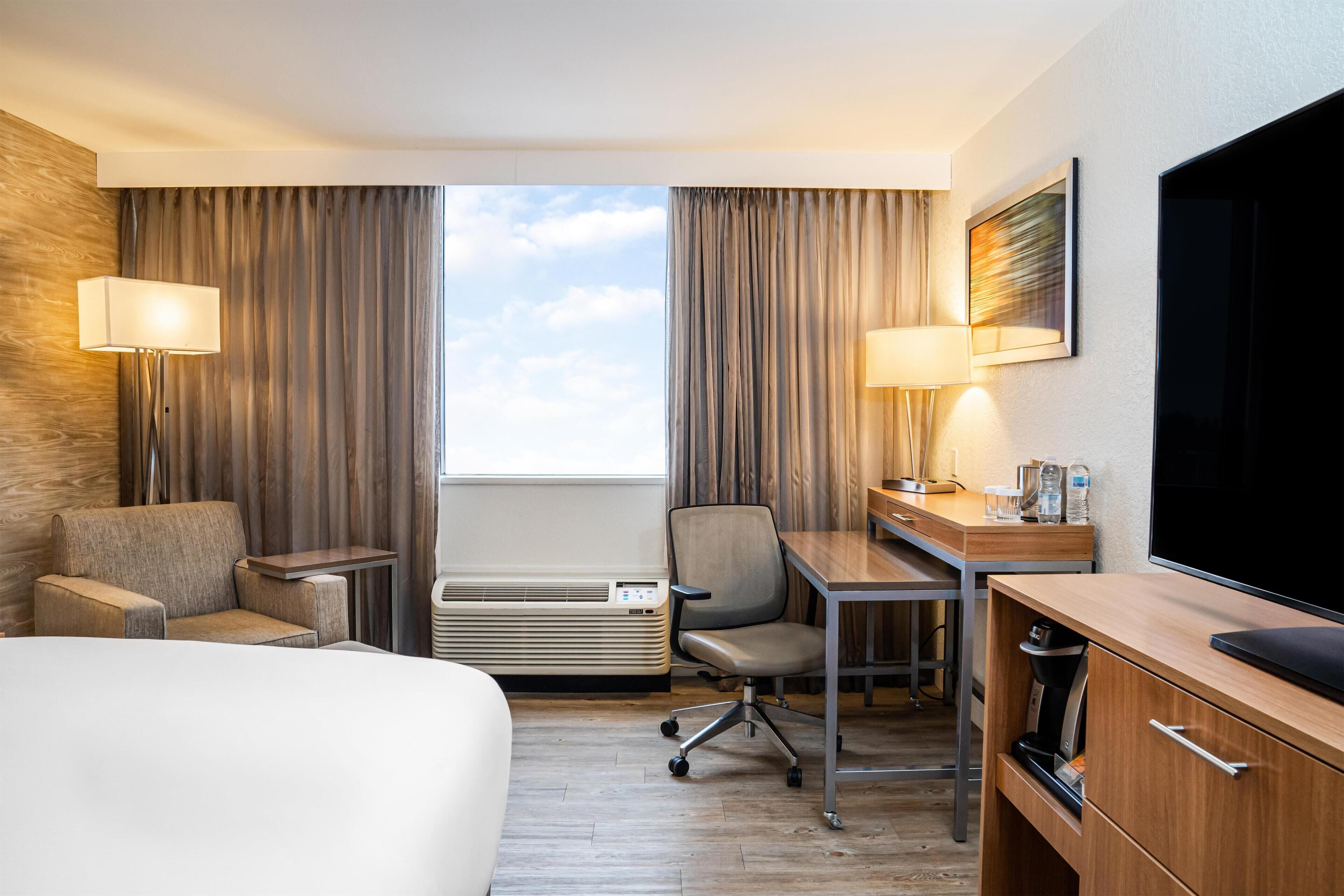 Quarto de hotel com cama, escrivaninha, cadeira, televisão, abajur e janela com cortinas.