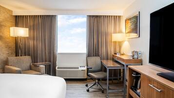 Quarto de hotel com cama, escrivaninha, cadeira, televisão, abajur e janela com cortinas.