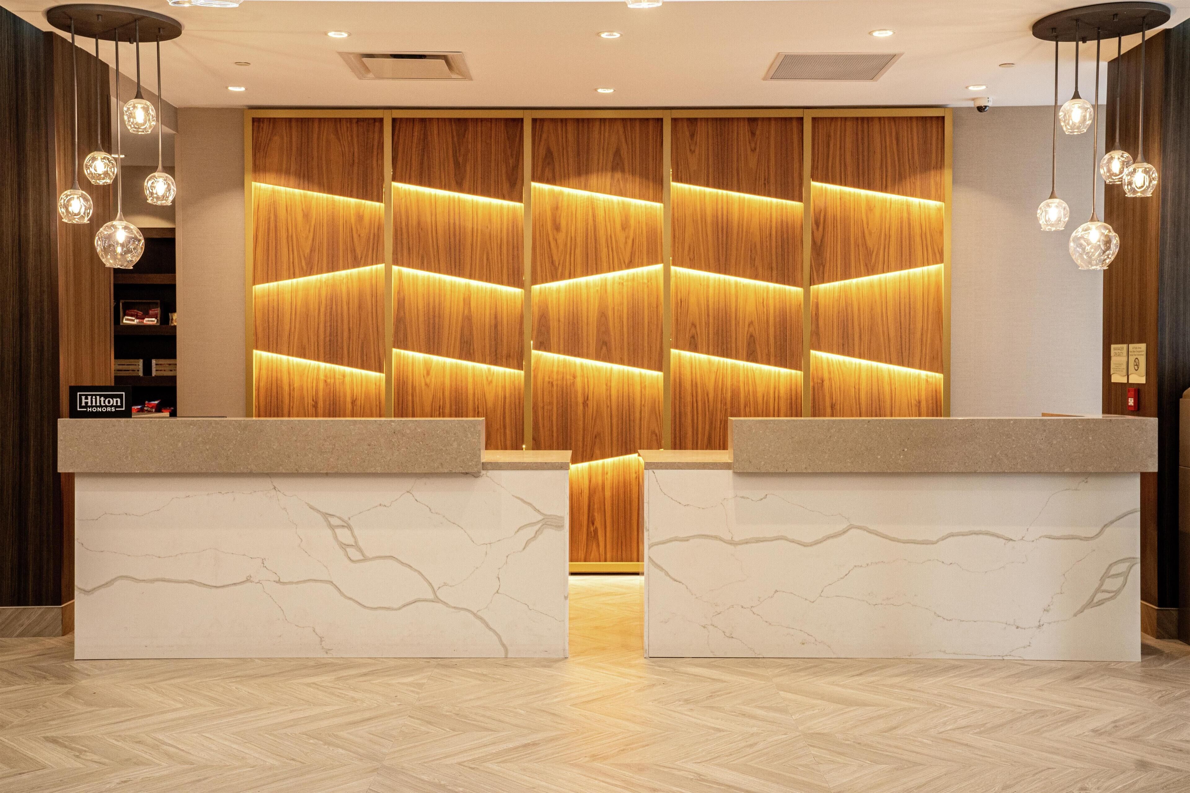 A hotel reception area with a wooden panel wall, marble reception counters, and hanging pendant lights.
