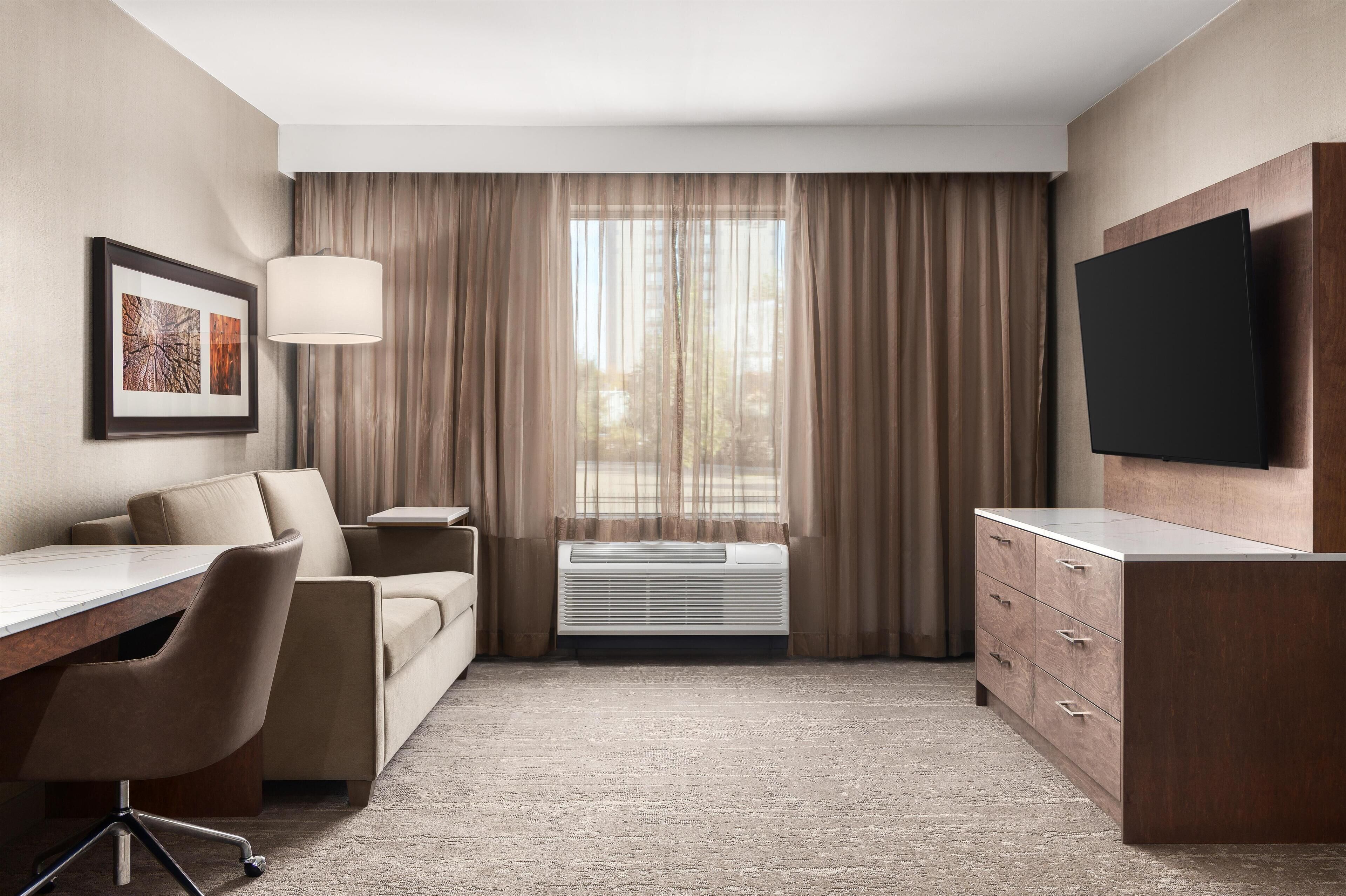 A hotel room with a desk, chair, sofa, air conditioning unit, TV, and a large window with curtains.