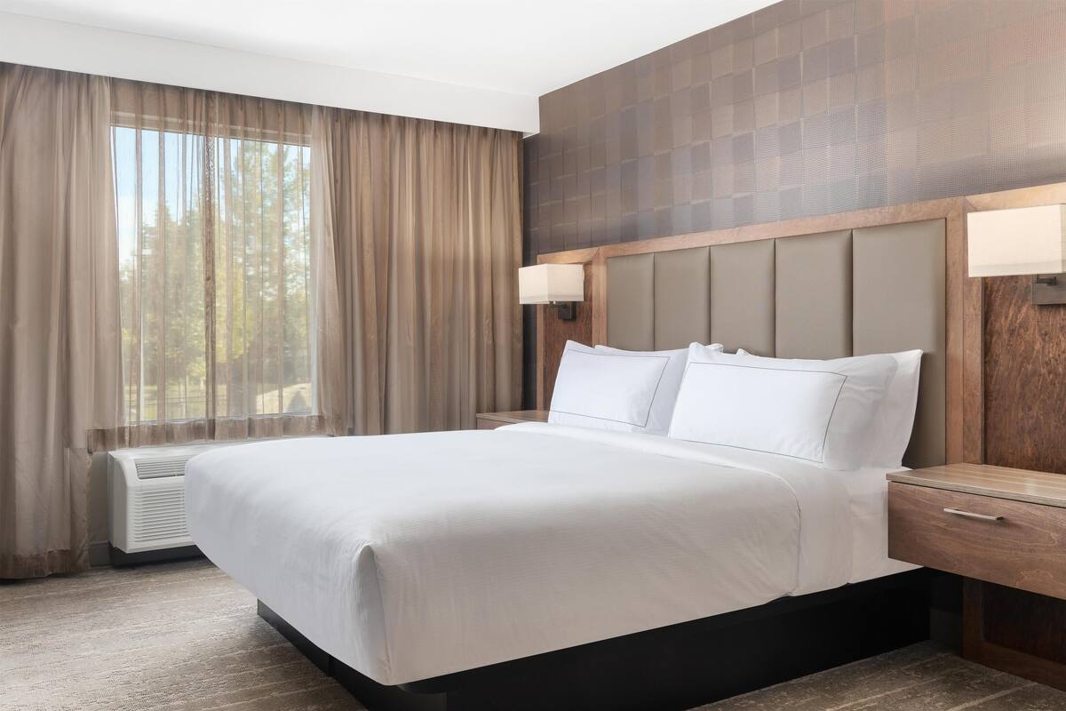 A hotel room with a large bed, a wooden headboard, a nightstand with a lamp, and a window with curtains.