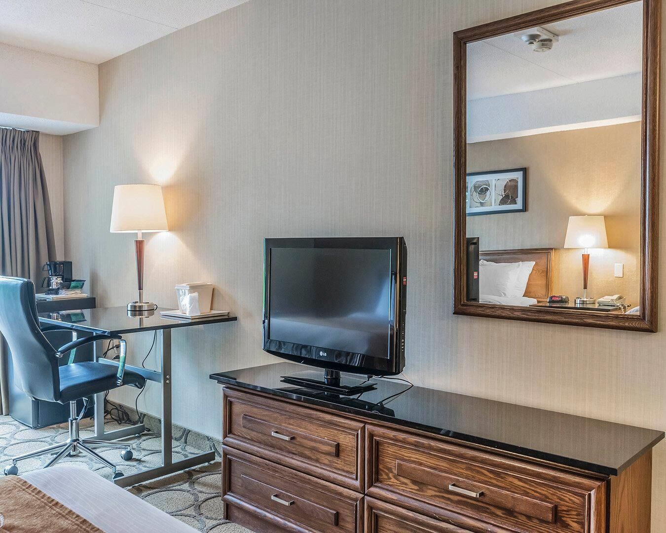 Quarto de hotel com televisão de tela plana, escrivaninha de madeira, cadeira, cômoda e um espelho grande.