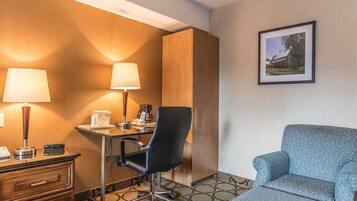A hotel room with a desk, two lamps, a chair, a sofa, a wooden wardrobe, and a framed picture on the wall.