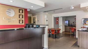 A hotel reception area with a wooden reception desk, a digital check-in station, and a kitchenette in the background.