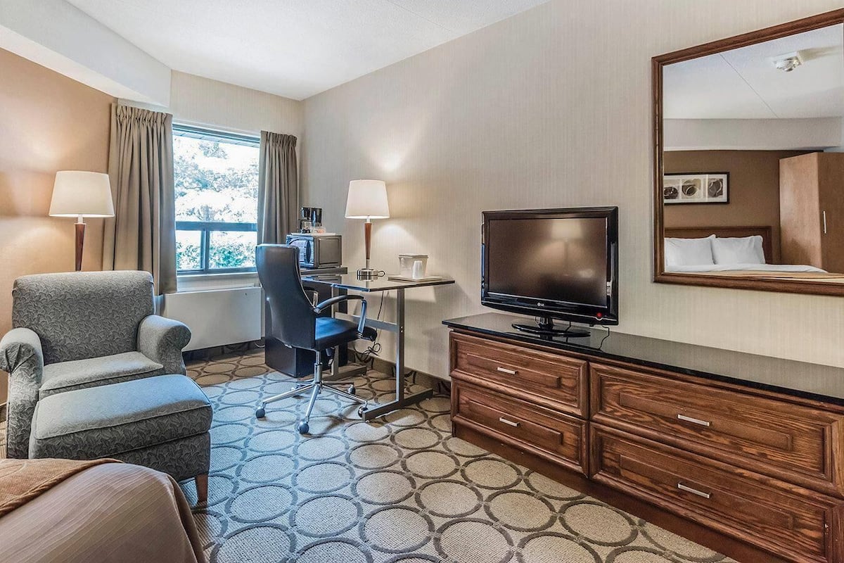 Une chambre d’hôtel comprenant un lit, un bureau avec un ordinateur, une télévision, une chaise, une commode et une fenêtre avec des rideaux.