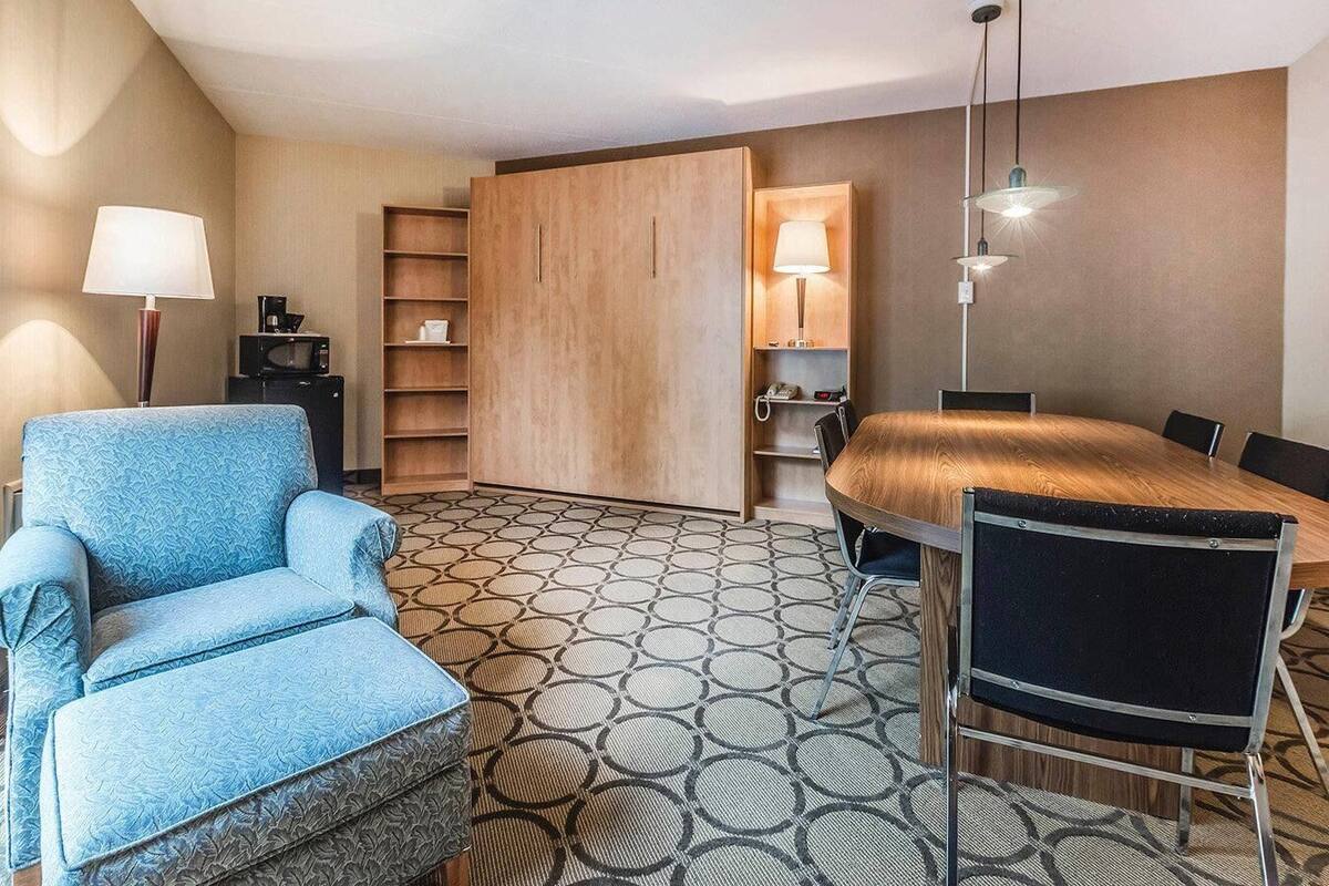 Une chambre d’hôtel avec un fauteuil bleu, une armoire en bois, une table à manger avec des chaises, un sol à motifs et une lampe.