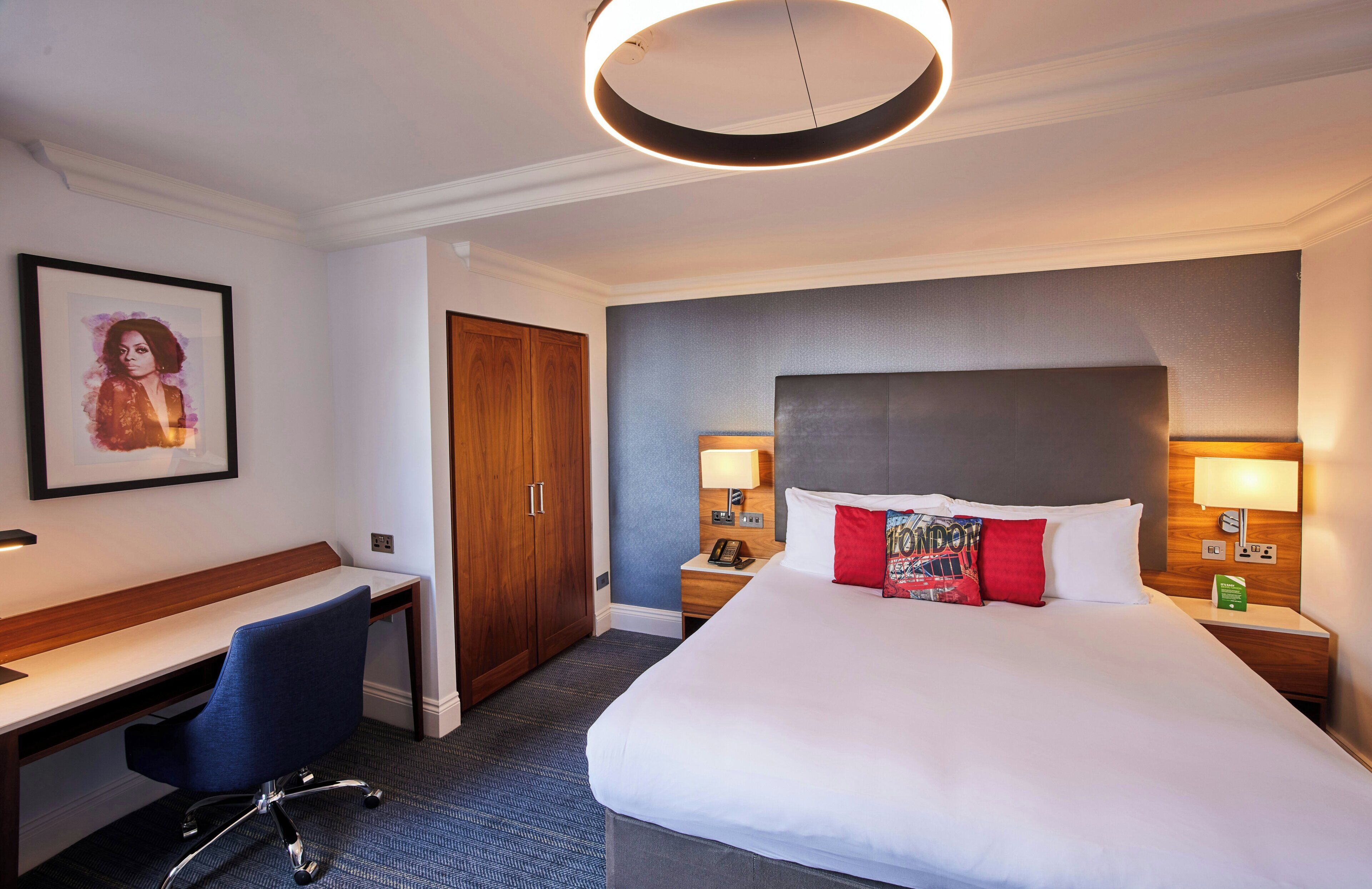 A hotel room with a large bed, a desk, a chair, a wardrobe, and a round ceiling light.
