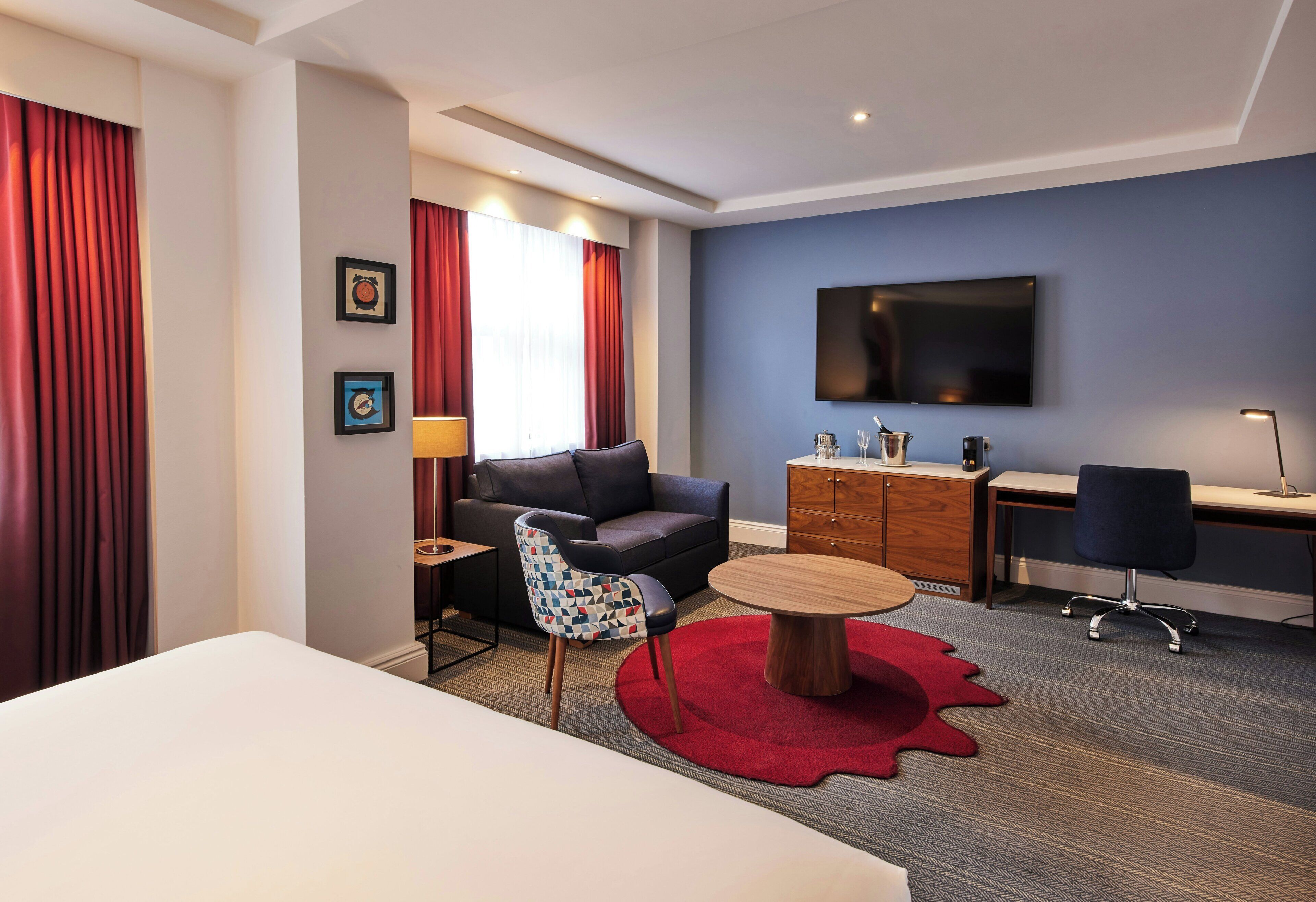 A hotel room with a bed, a desk, a chair, a sofa, a TV, and a red rug.