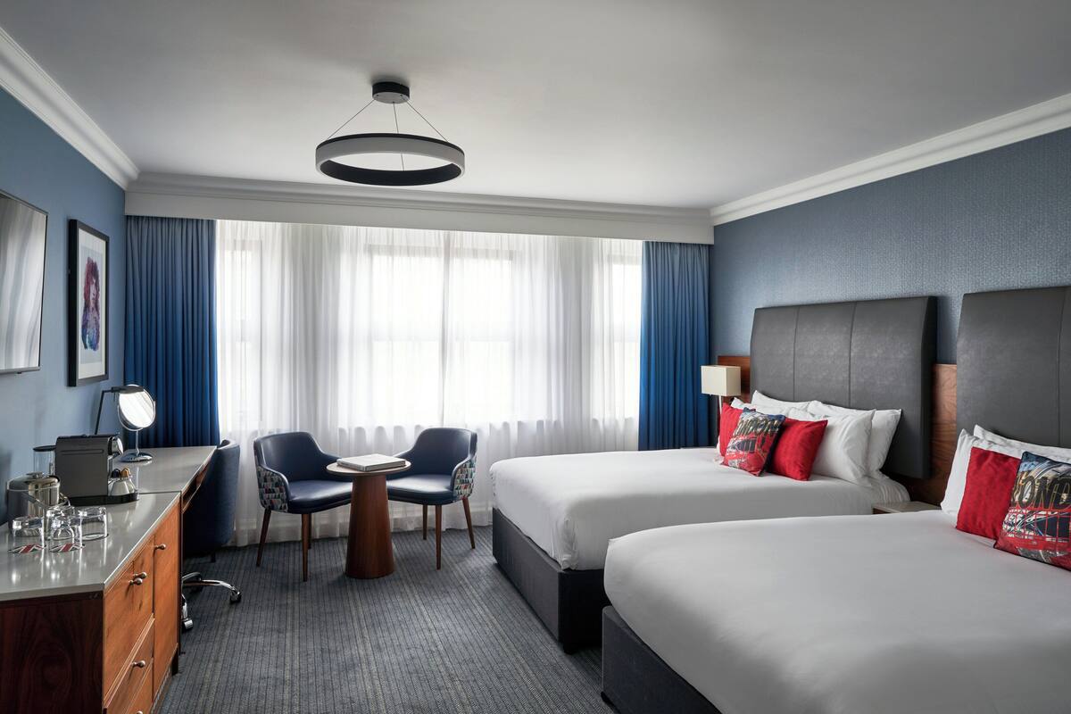 Hotellihuone, jossa on kaksi sänkyä, työpöytä, tuoli, työpöytälamppu ja ikkuna, jossa on verhot.