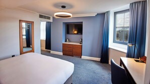 A hotel room with a large bed, a desk, a dresser, a mirror, a television, and a window with curtains. - The Cumberland, London (London)