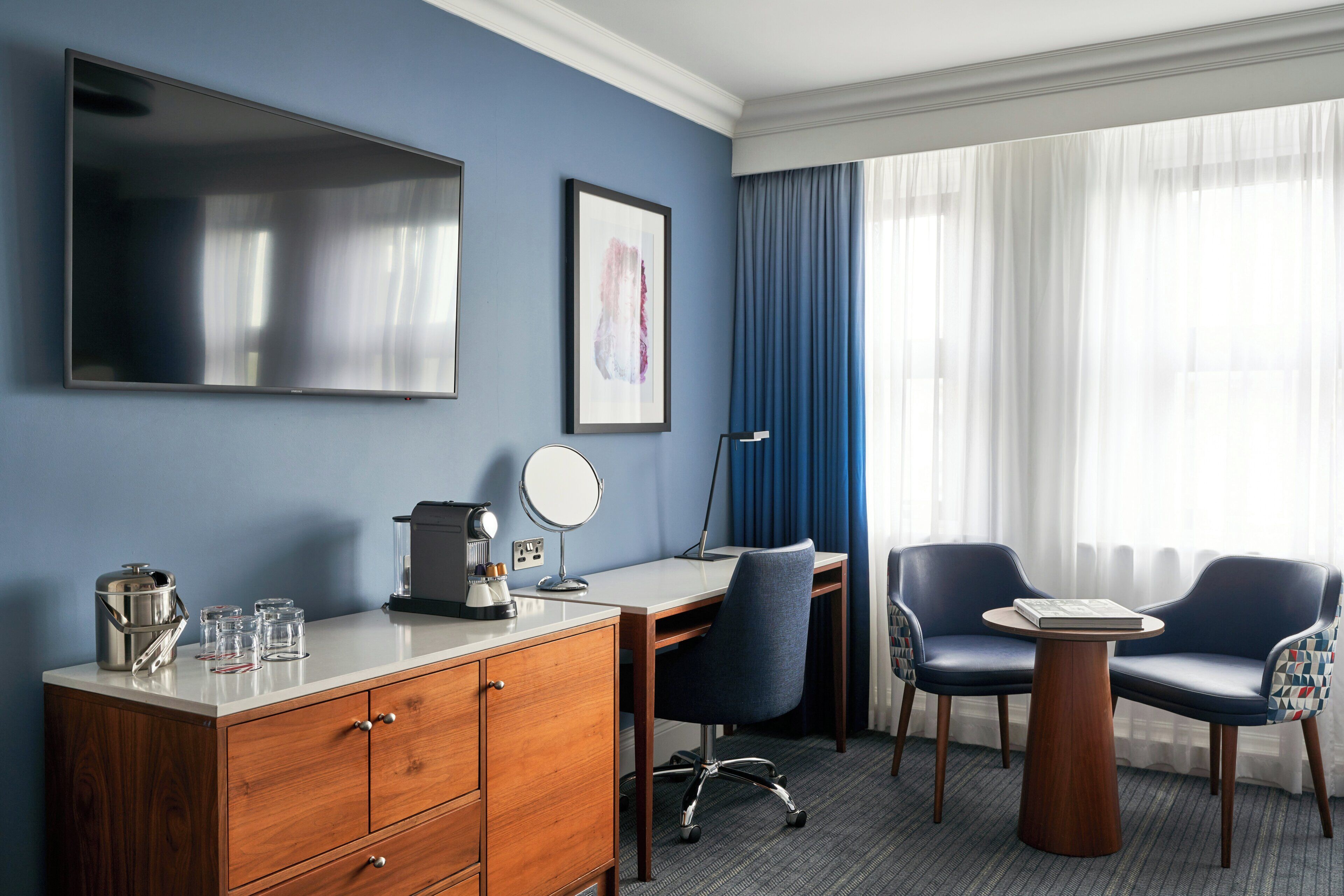 A hotel room with a desk, two chairs, a coffee maker, a lamp, a TV, and a window with curtains.