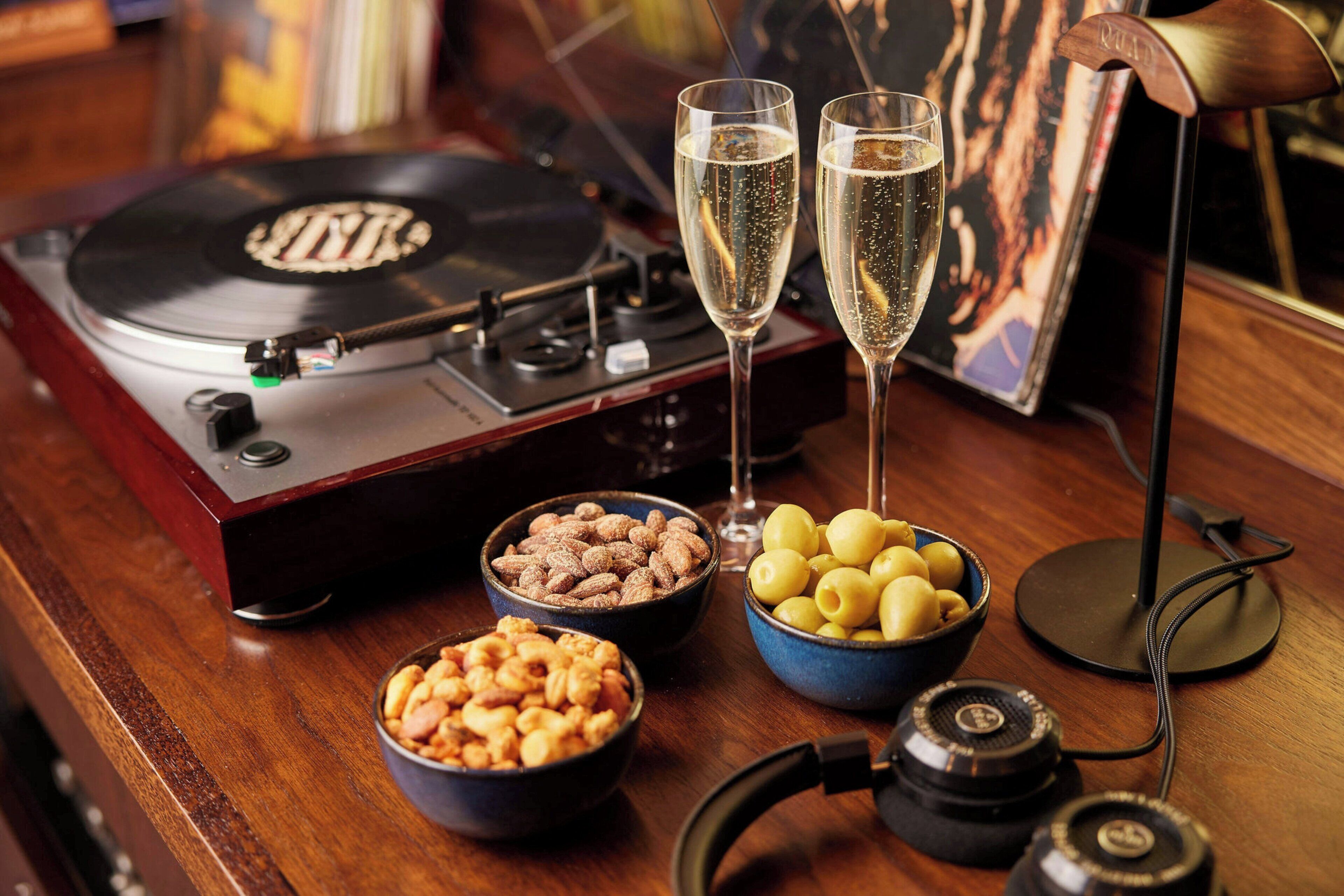 Tocadiscos, dos copas de champán, cuencos con frutos secos y aceitunas, y un par de auriculares sobre una mesa de madera.