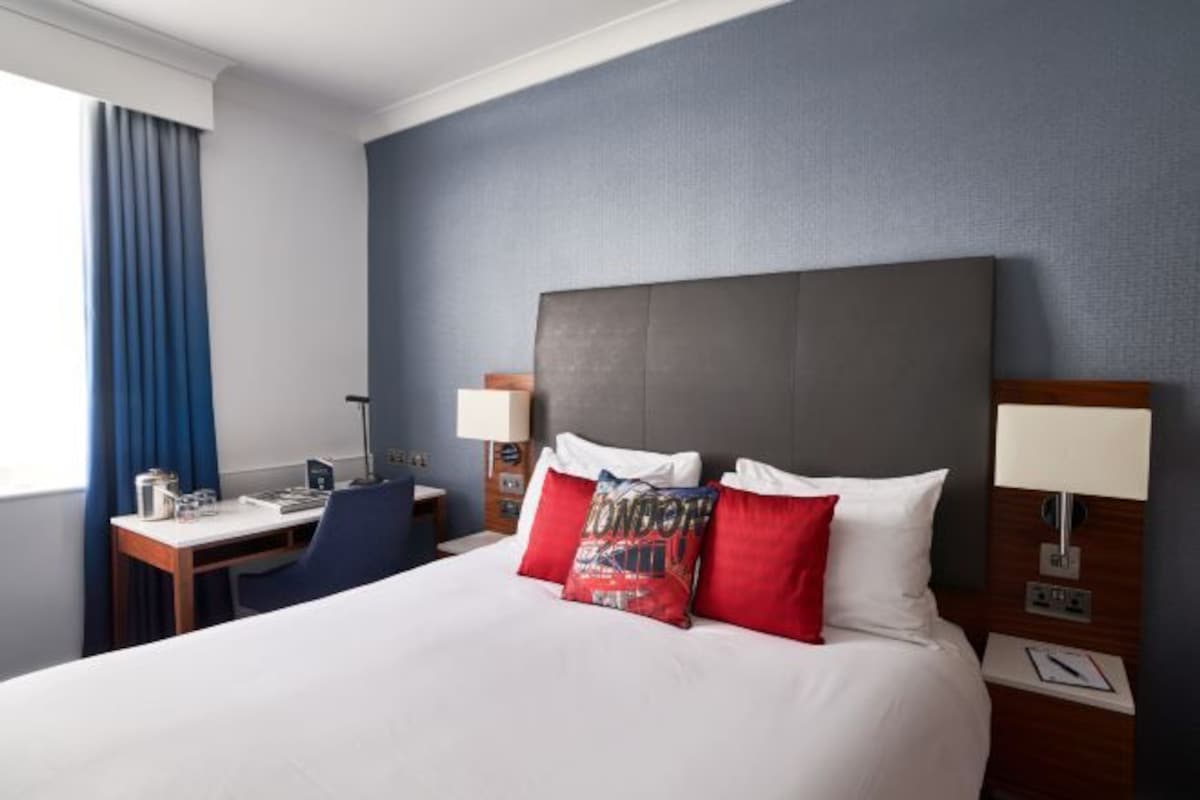 A hotel room with a large bed, a desk, a chair, and a nightstand with a lamp.