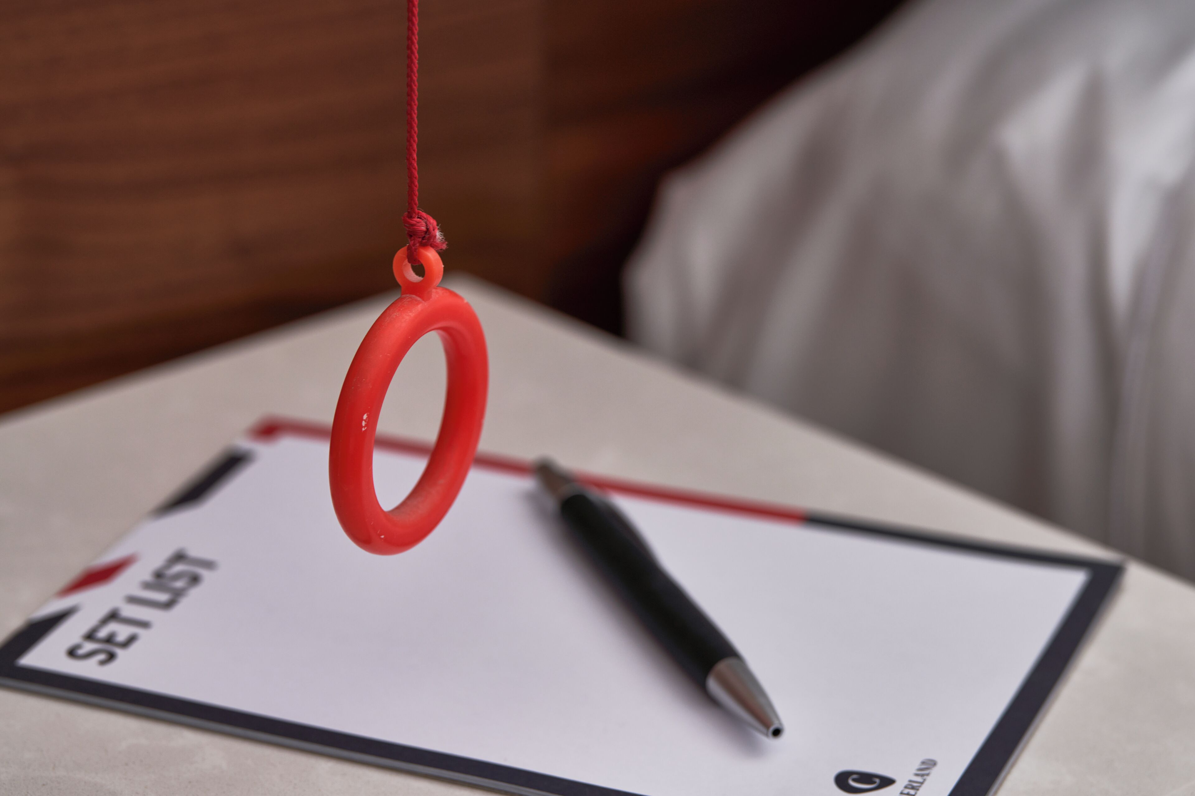 Ein roter, kreisförmiger Gegenstand hängt an einer Schnur, ein schwarzer Stift und ein Dokument mit dem Schriftzug "SET LIST" sowie einem Logo befinden sich auf einem Tisch.