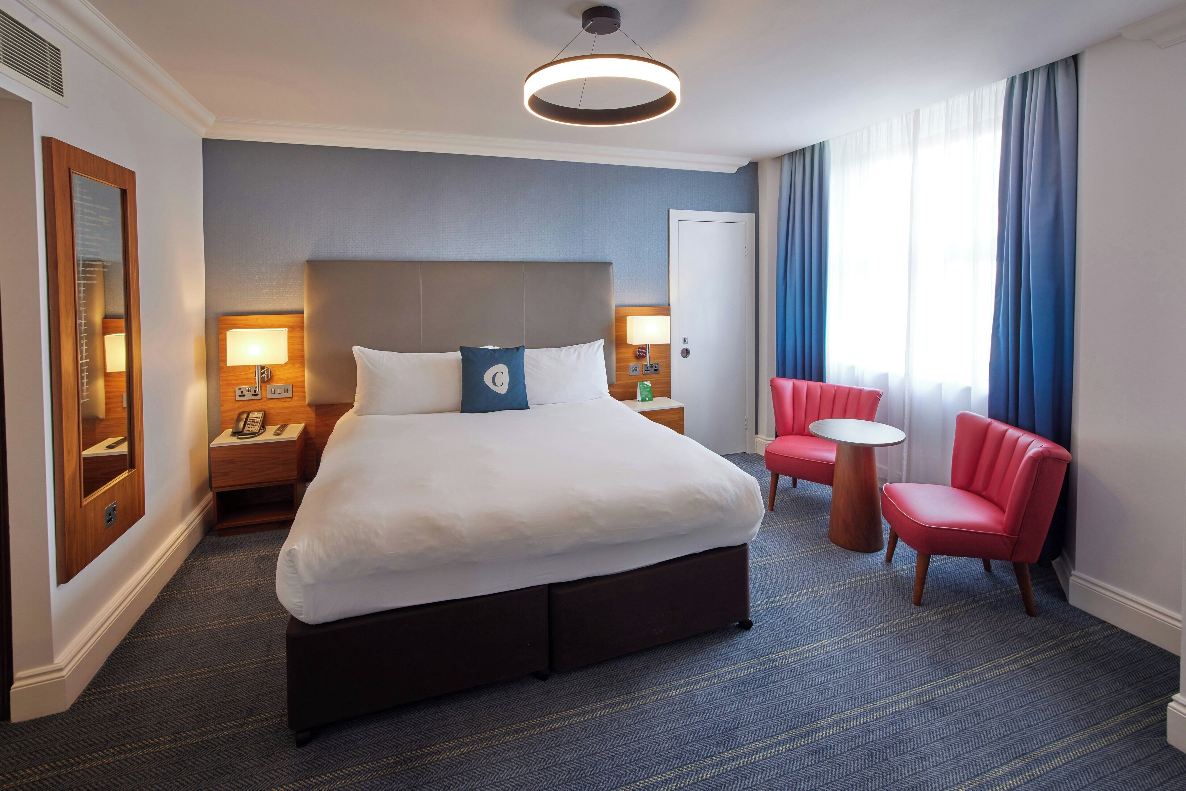 Una camera d'albergo con un letto grande, due comodini con lampade, un tavolino rotondo, una sedia rossa e un'ampia finestra con tende.