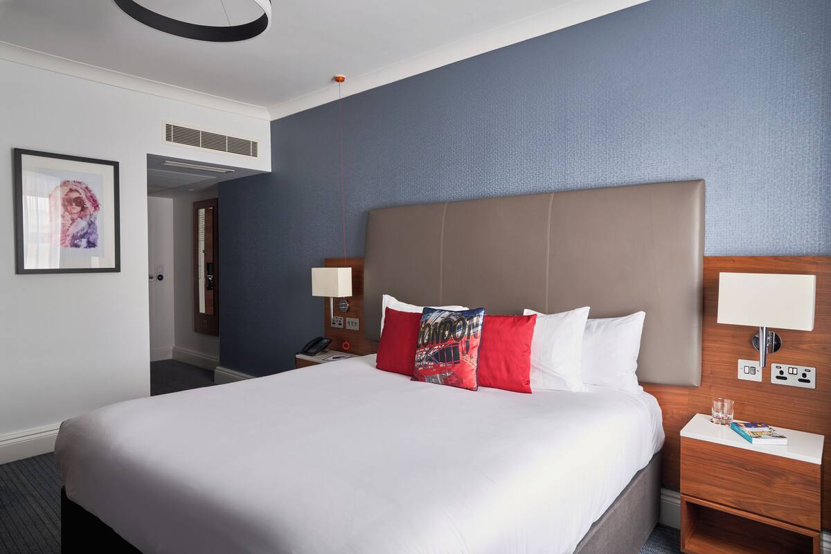 Ein Hotelzimmer mit einem großen Bett, zwei Nachttischen, einem Beistelltisch, einem gerahmten Bild an der Wand und einer Tür zu einem anderen Raum.