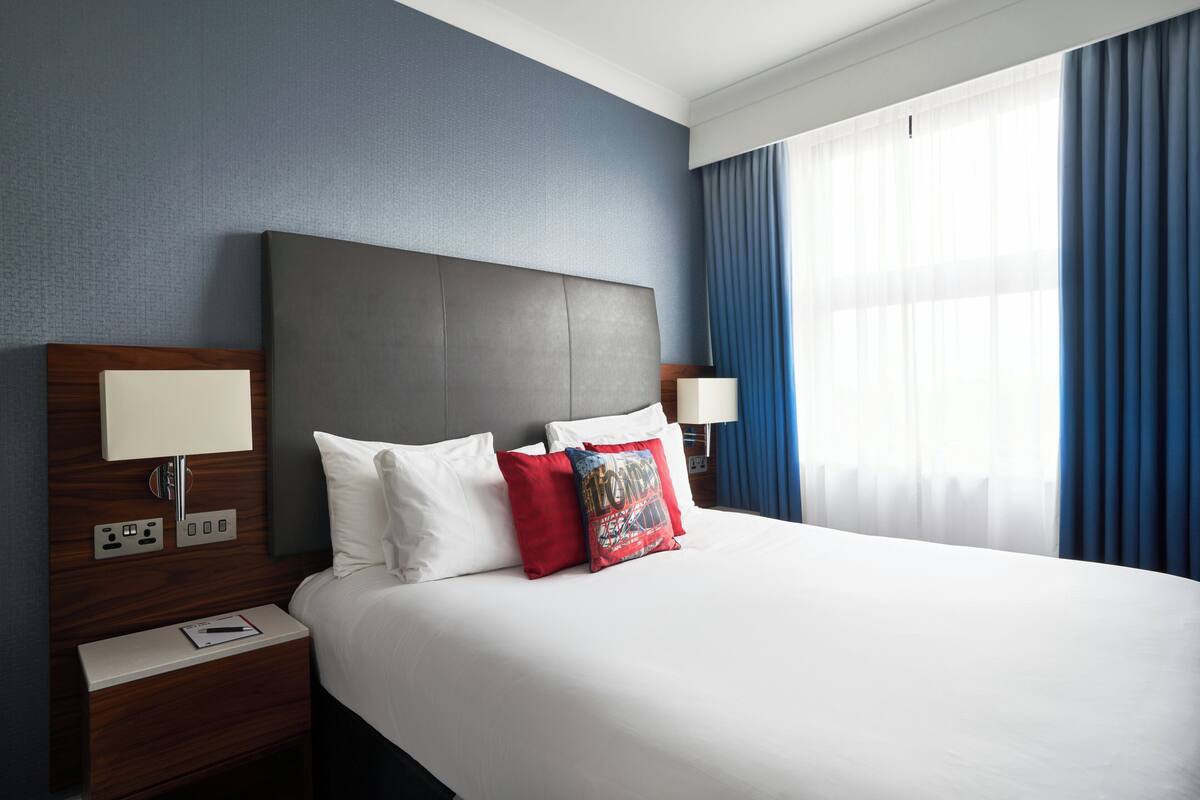 Ein Hotelzimmer mit einem großen Bett, einem Nachttisch mit Lampe und einem Fenster mit Vorhängen.