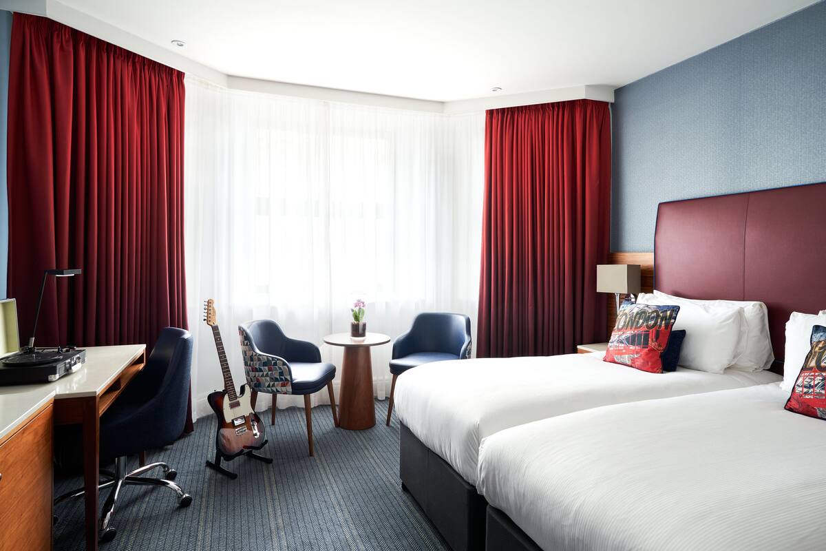 Ein Hotelzimmer mit zwei Betten, einem Schreibtisch mit Computer, einem Stuhl, einer Gitarre und einem kleinen Tisch mit Pflanze.