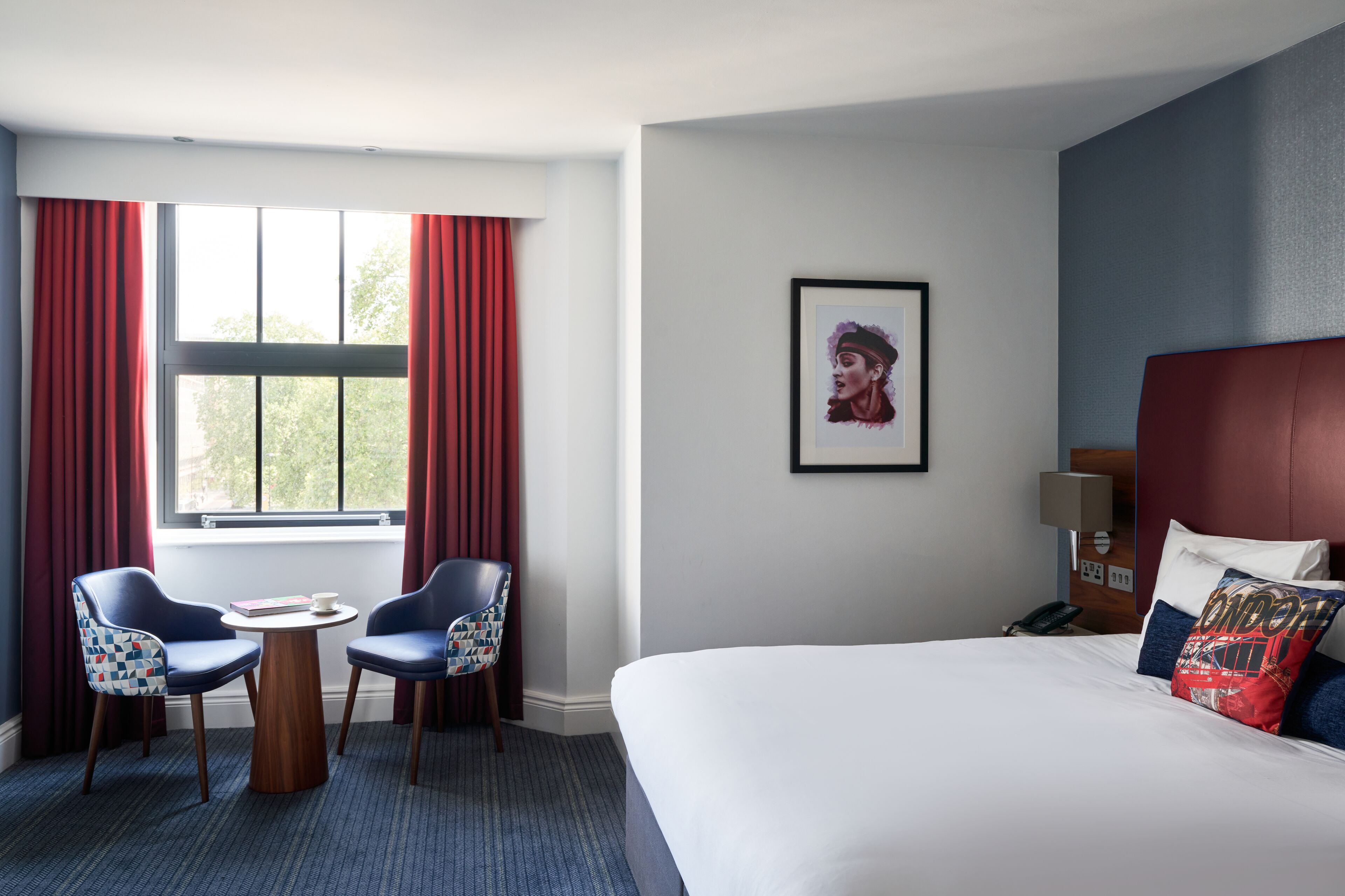 A hotel room with a bed, two chairs, a small table, a window with red curtains, a framed picture on the wall, and a nightstand with a lamp.