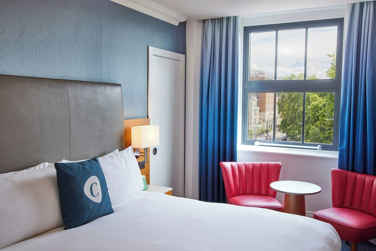 Hotellihuone, jossa on sänky, yöpöytä, lamppu, ikkuna verhoilla ja kaksi punaista tuolia.