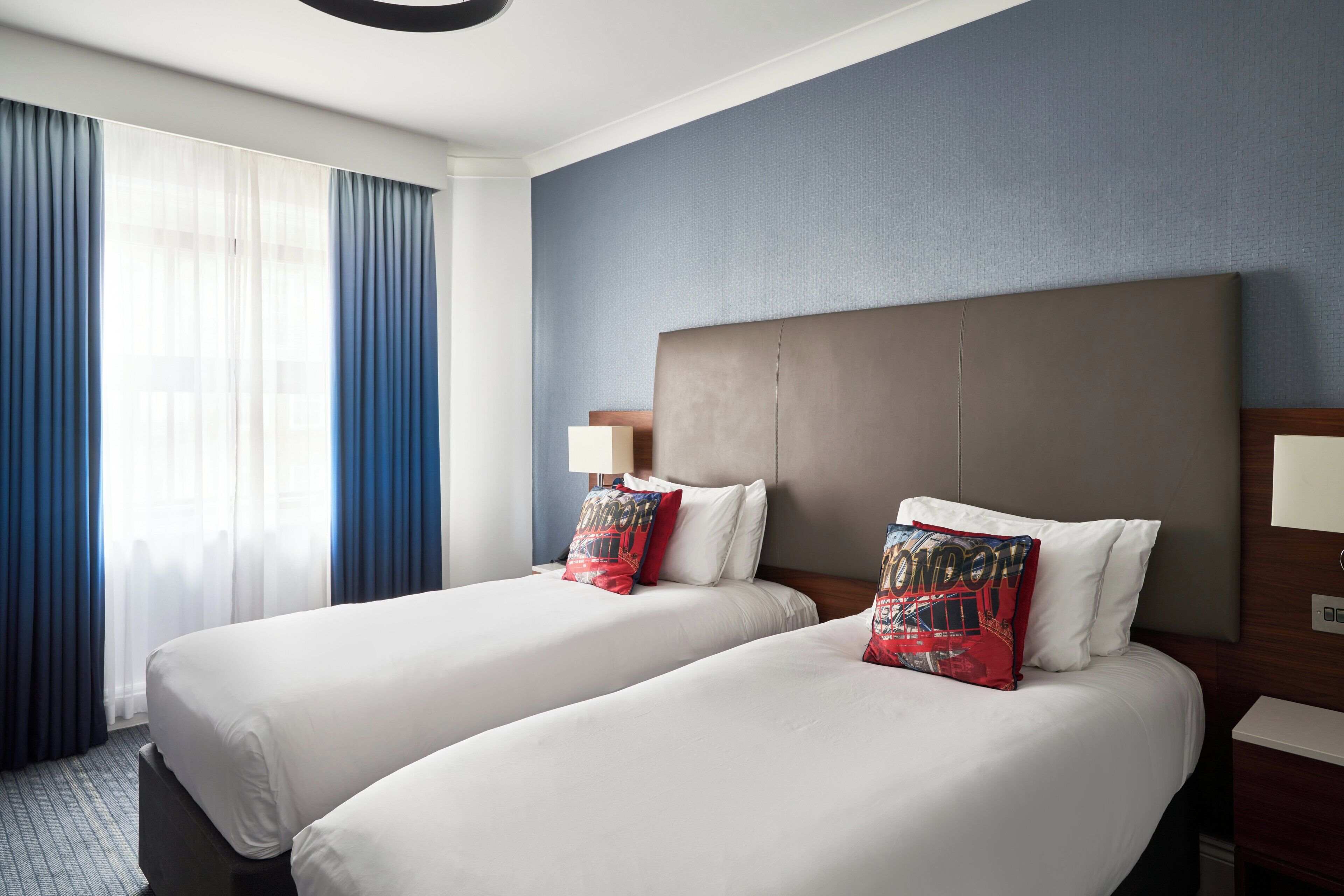 Ein Hotelzimmer mit zwei Betten, einem großen Fenster mit blauen Vorhängen und einer blauen Wand.