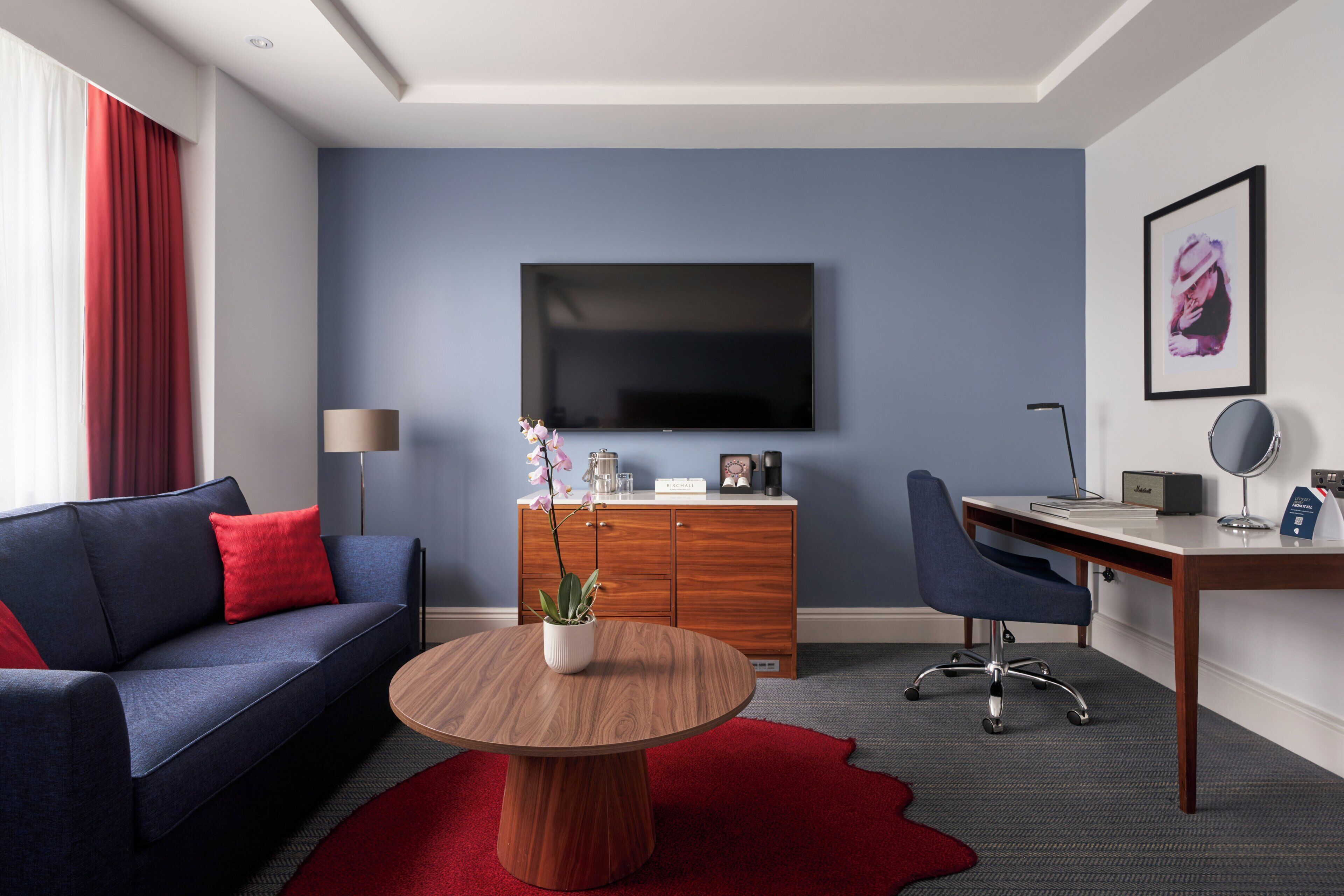 A modern hotel room with a blue sofa, a round wooden coffee table, a desk with a chair, a flat-screen TV, and a cabinet.