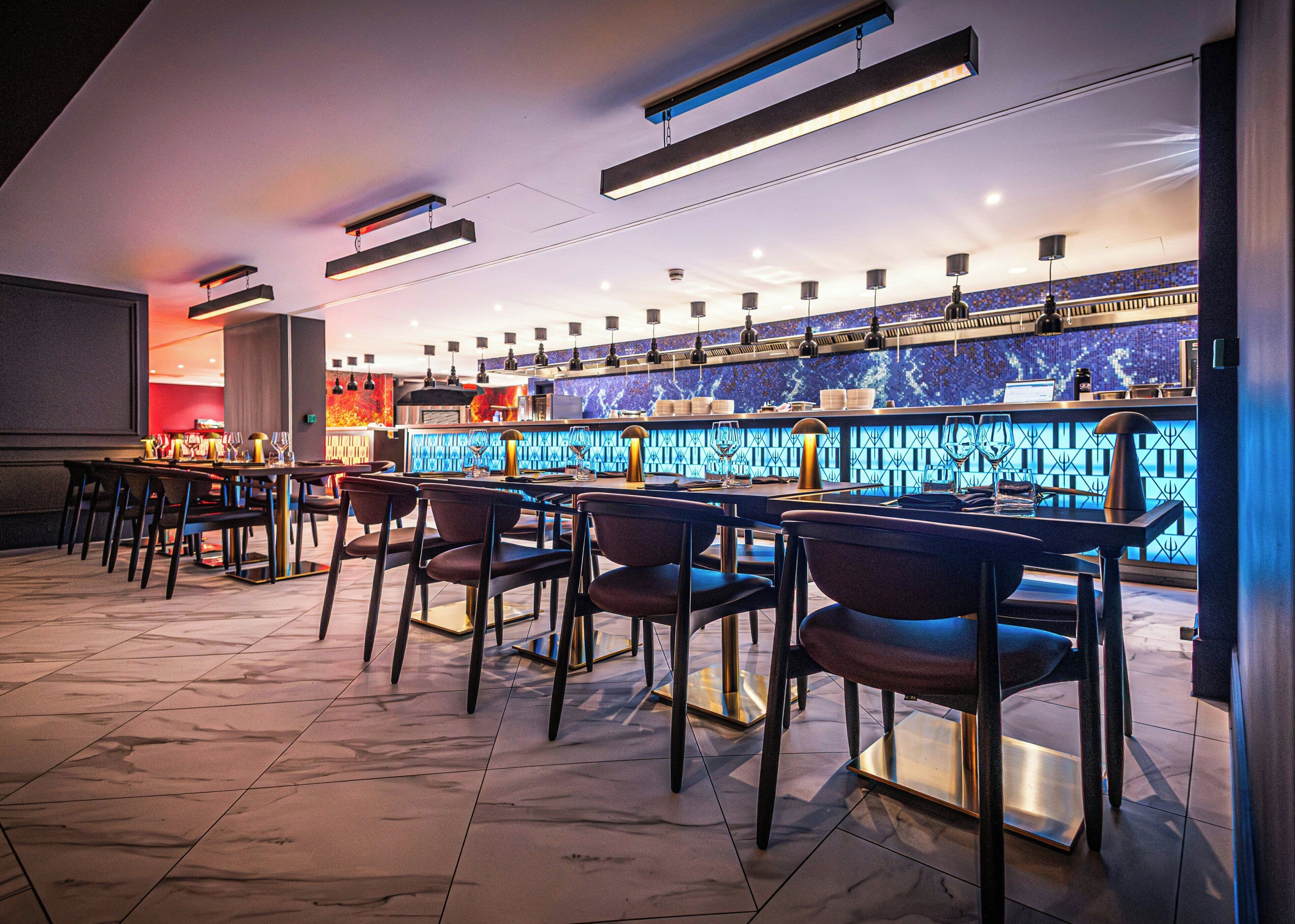 A bar area with multiple tables and chairs, a counter with a blue patterned backsplash, and overhead lighting fixtures.
