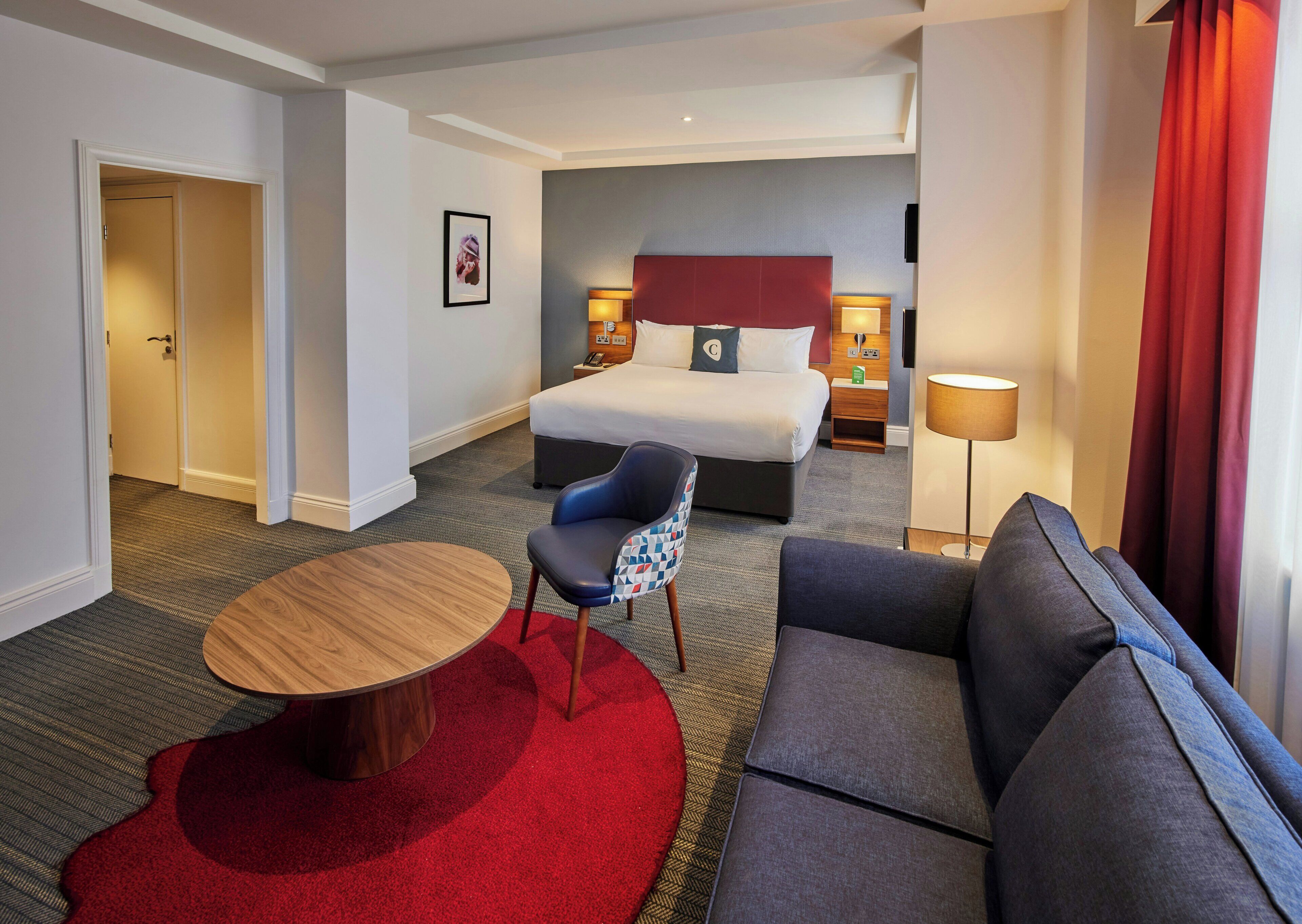 A hotel room with a bed, a sofa, a chair, a small table, and a lamp.