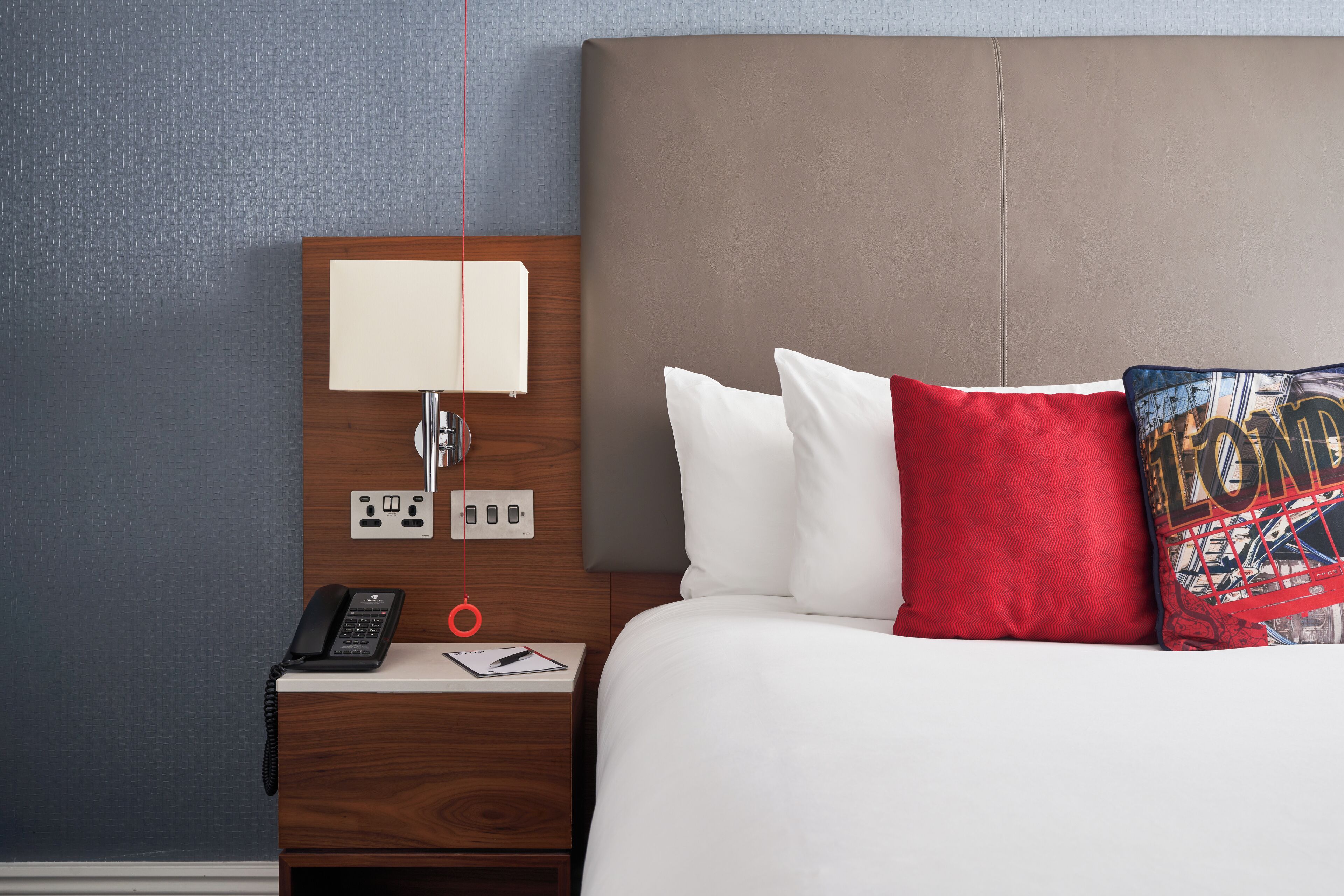 A hotel room with a wooden nightstand, a lamp, a phone, a notepad, and a bed with white and red pillows.