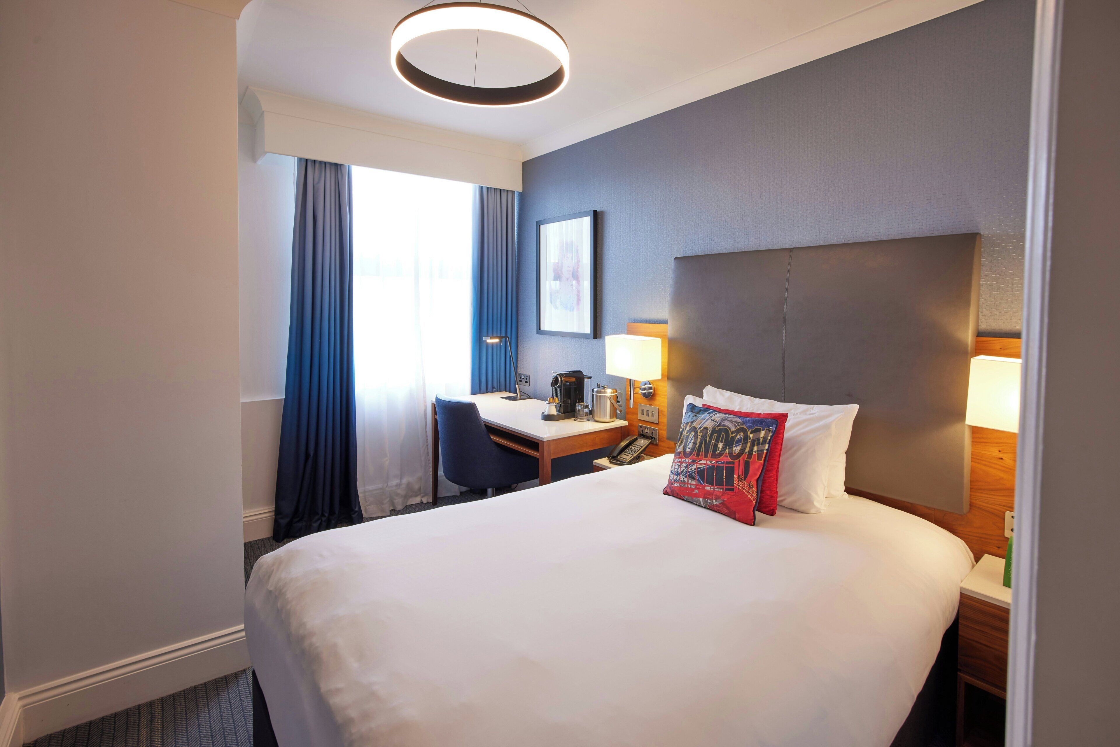 A hotel room with a large bed, a desk with a chair, a lamp, and a window with curtains.