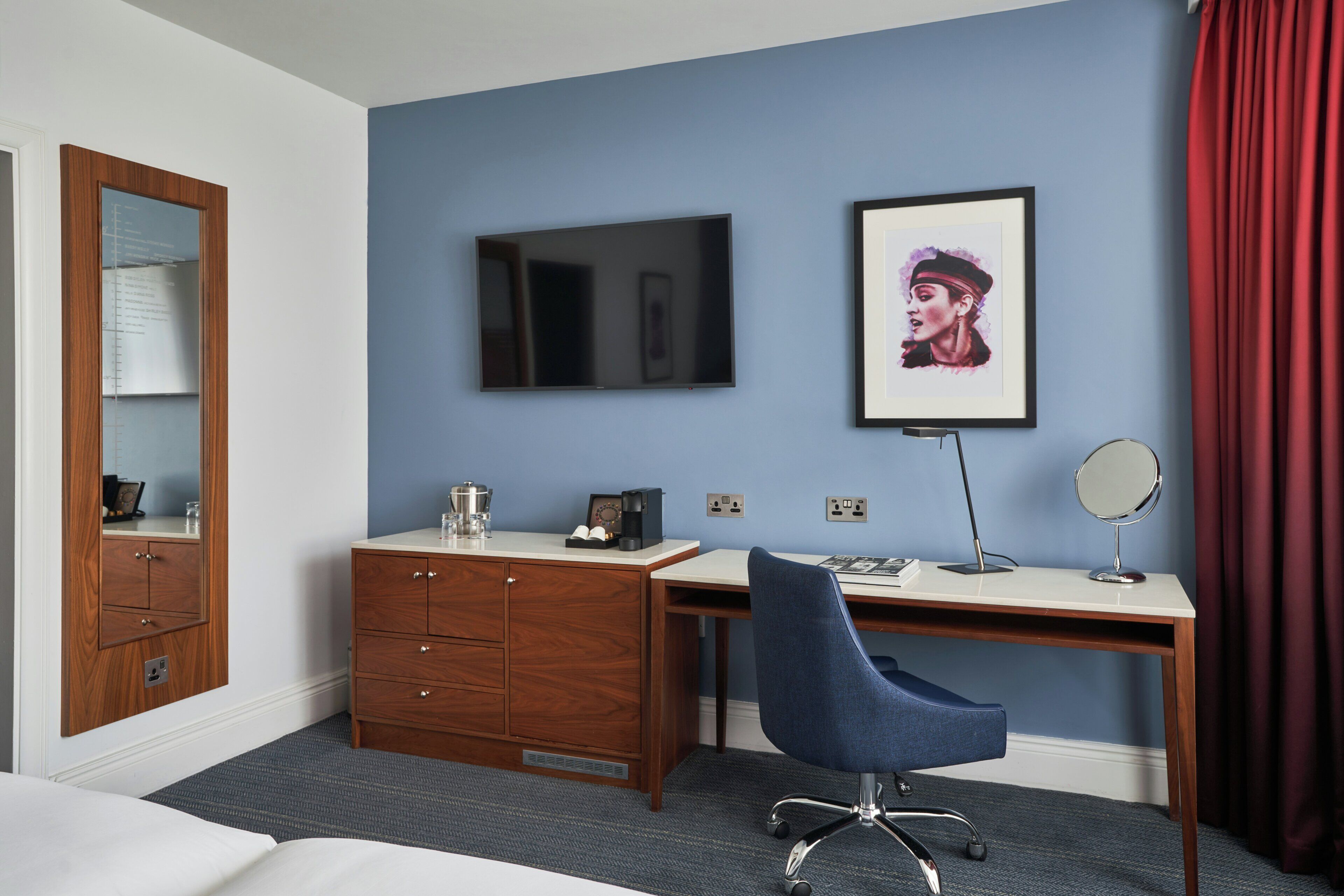 Ein Hotelzimmer mit Bett, Schreibtisch mit Stuhl, Kommode, Spiegel und einem gerahmten Kunstwerk an der Wand.