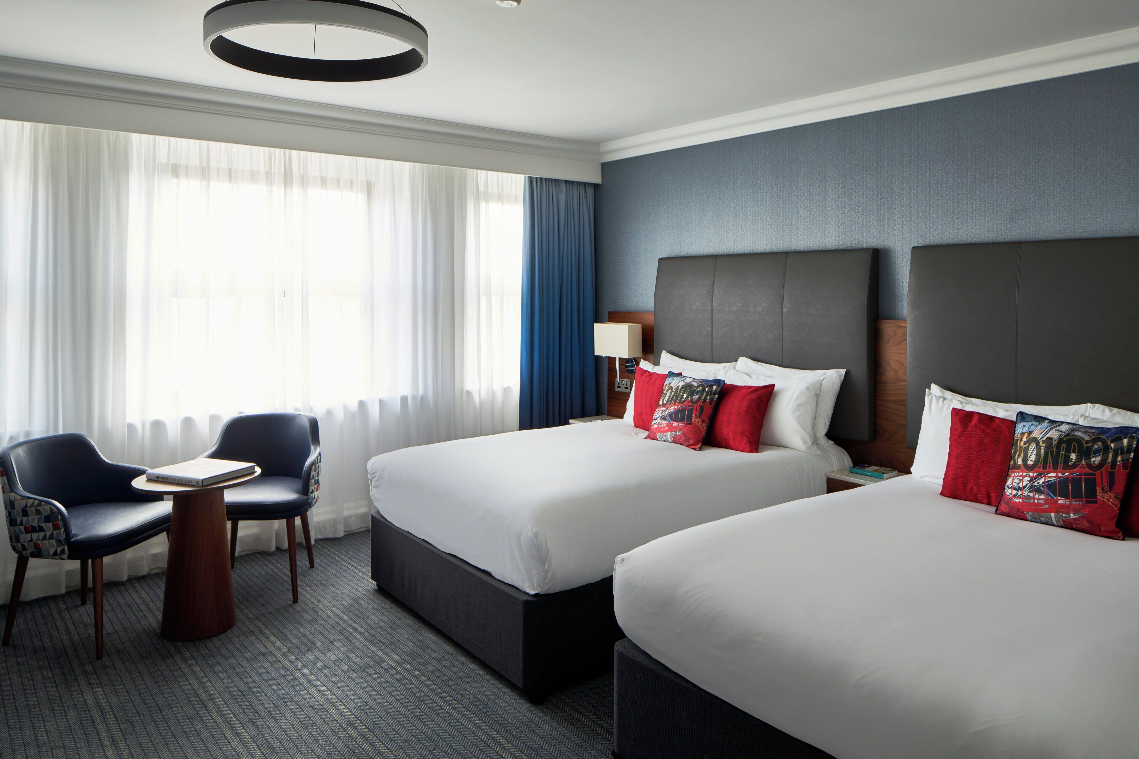 A hotel room with two beds, a desk, a chair, and a large window with curtains.