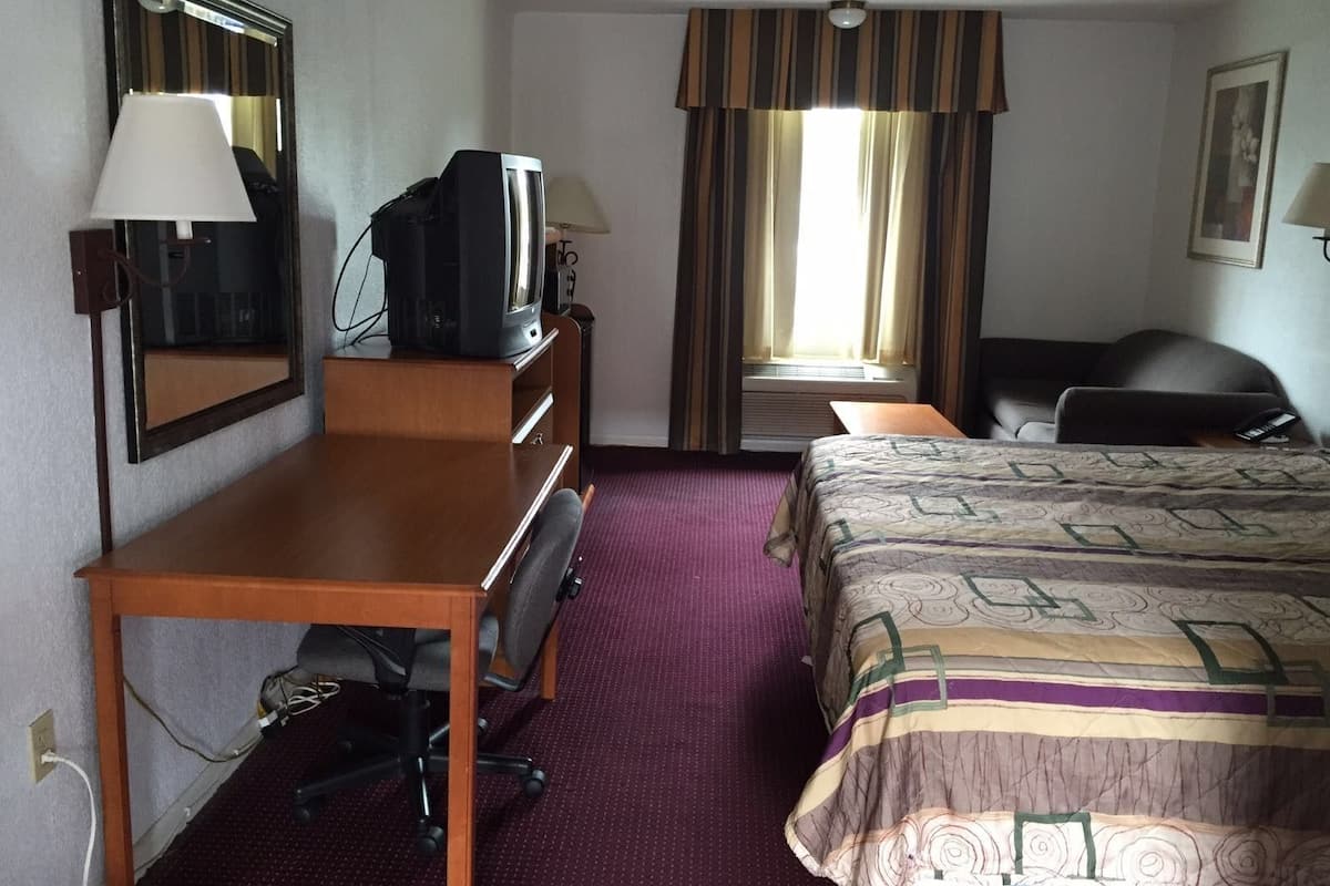 A hotel room with a bed, a desk with a chair, a television, a mirror, and a window with curtains.