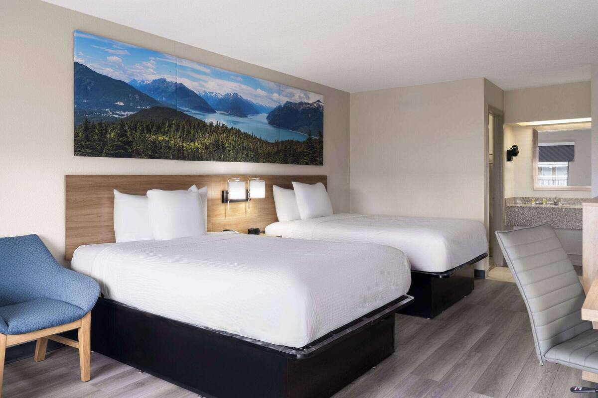 Een hotelkamer met twee bedden, een bureau, een stoel, een schilderij van een berglandschap en een kleine kitchenette.