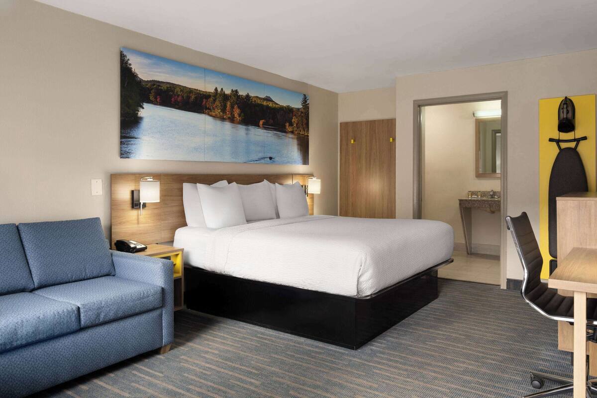Een hotelkamer met een bed, een blauwe bank, een bureau met een stoel, een schilderij van een meer en een spiegel aan de muur.